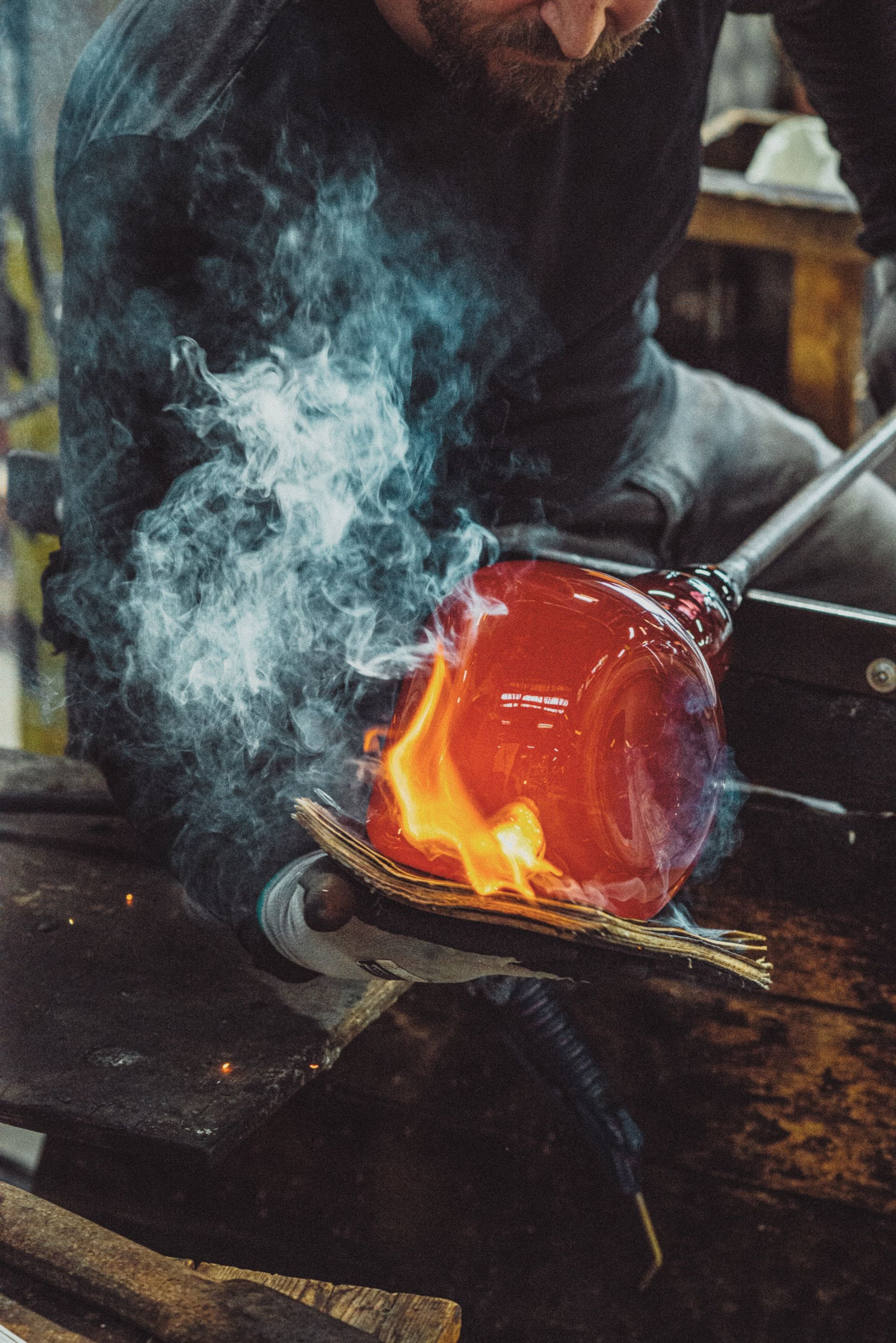 A glassblower working with molten red glass, flames and smoke visible in a workshop.