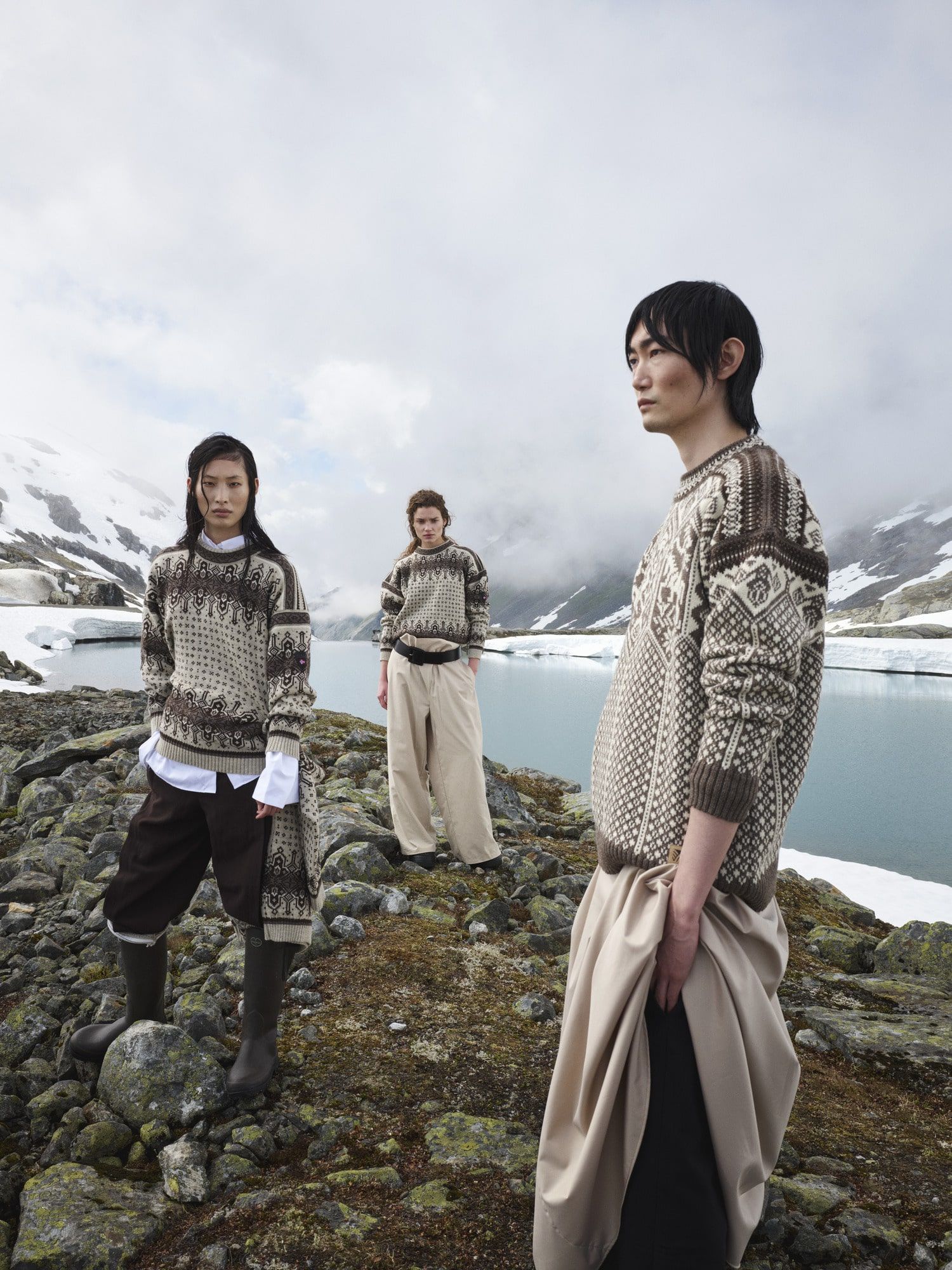 Three people in patterned sweaters stand by a body of water, snowy mountains in background.