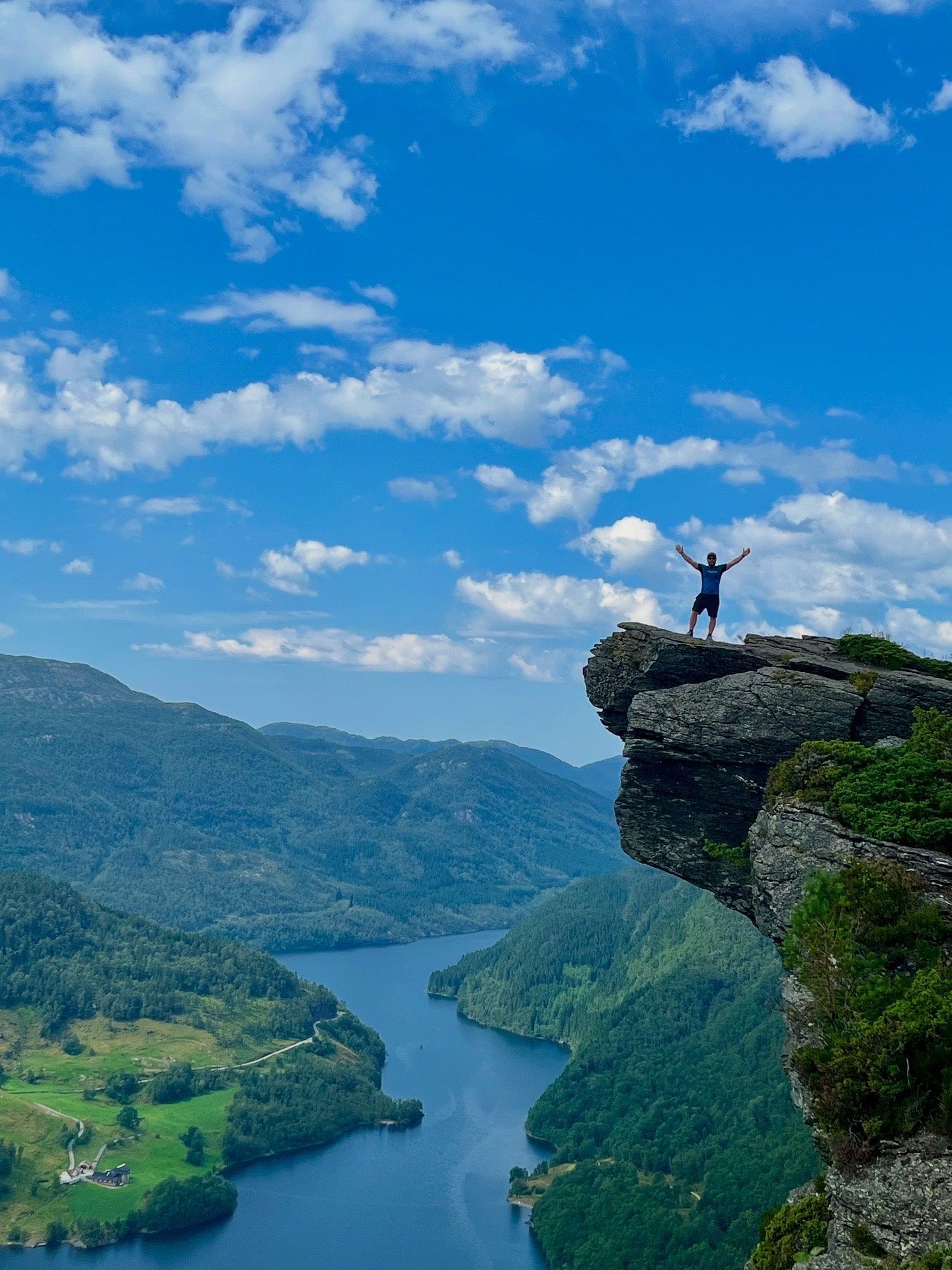 Person with arms raised on a cliff overlooking a fjord, blue sky with clouds.