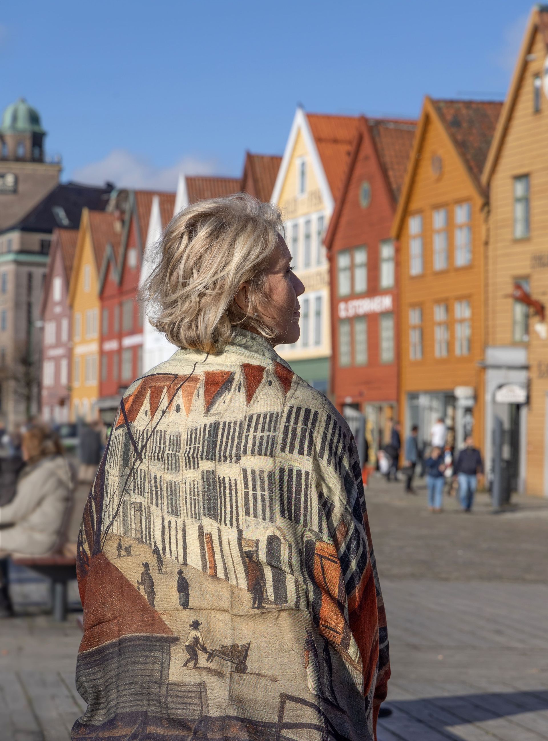 Woman wearing patterned jacket, looking towards colorful buildings in Bergen, Norway.
