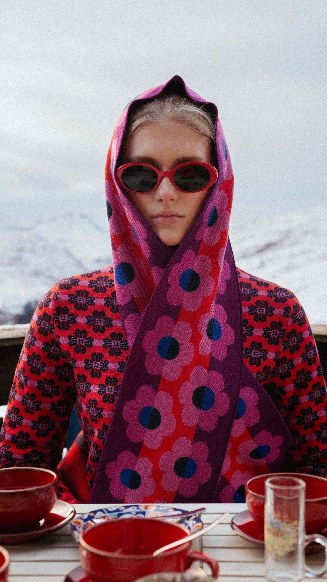 Woman wearing floral scarf and sunglasses, seated at outdoor table with red cups, snow-capped mountains in background.