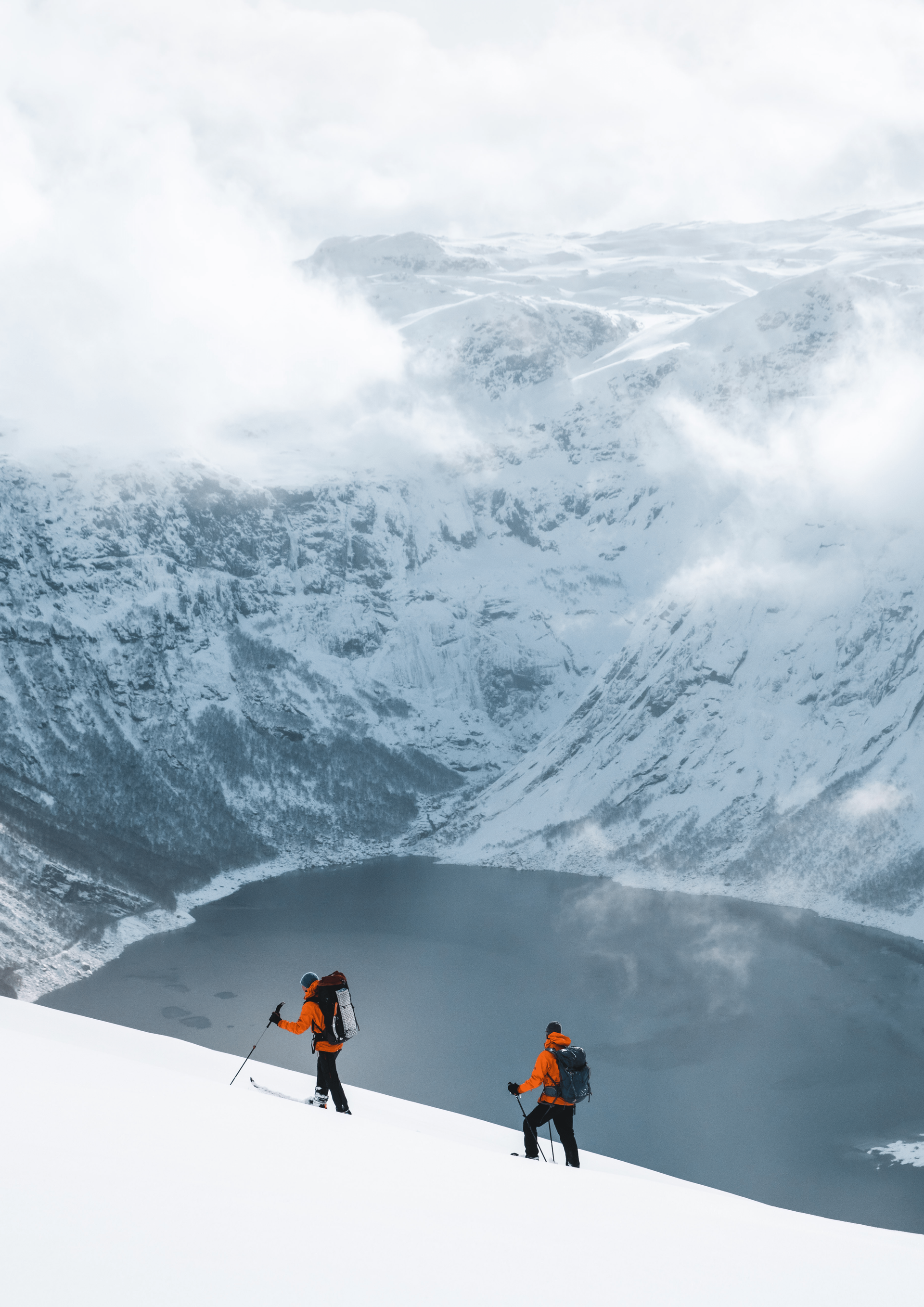 Two skiers ascend a snowy mountain, overlooking a dark lake, cloudy sky.