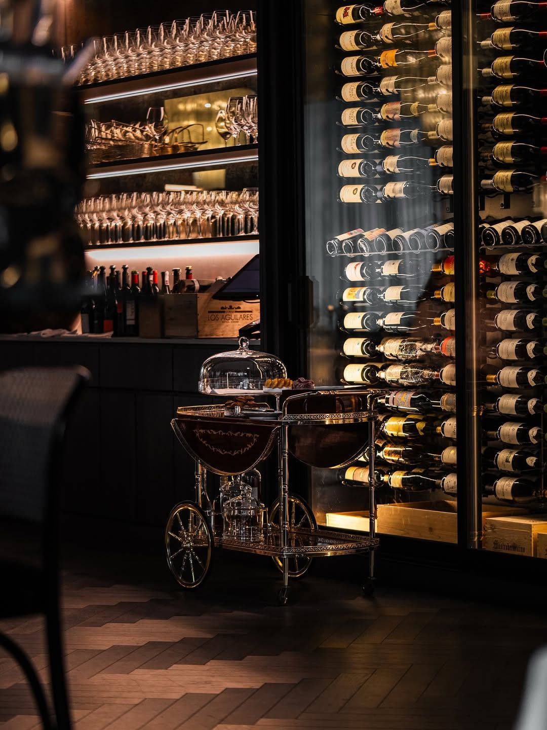 Wine cellar interior with wine cart, shelves of glasses, and rows of wine bottles.