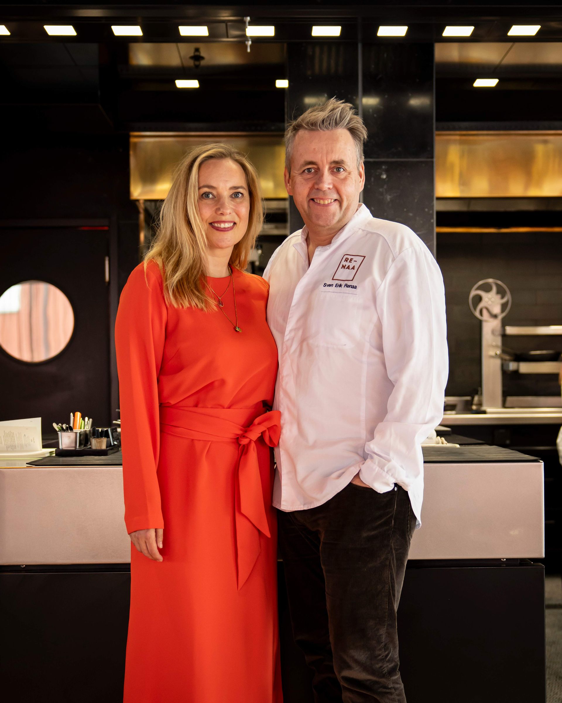 Woman in red dress and man in chef's coat pose together in a restaurant kitchen.