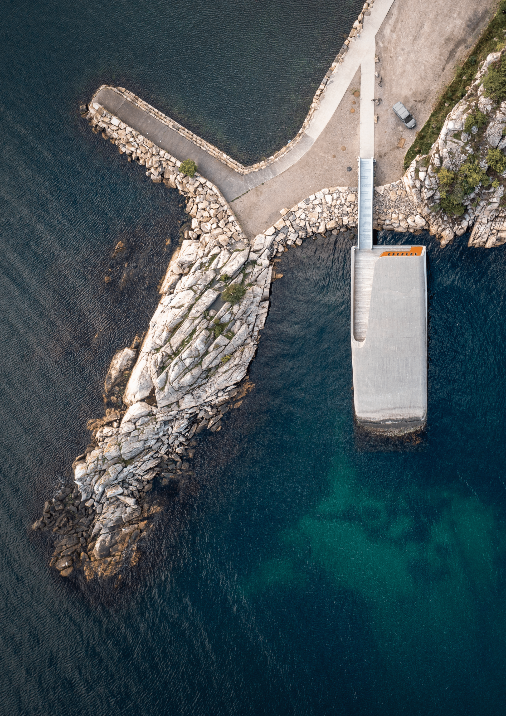 Aerial view of a pier and rocky outcropping extending into blue-green ocean water.