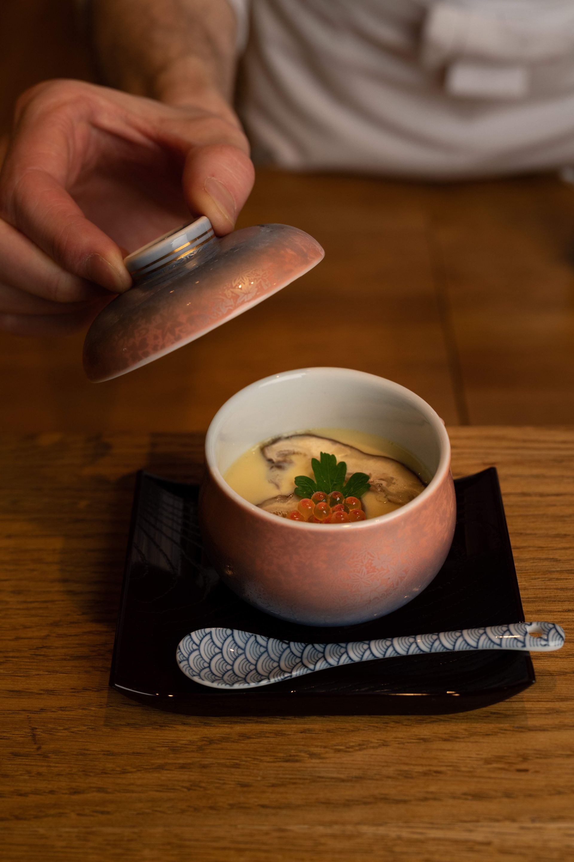 A hand lifts the lid off a bowl of Japanese chawanmushi, set on a black plate, with a spoon on a wooden table.