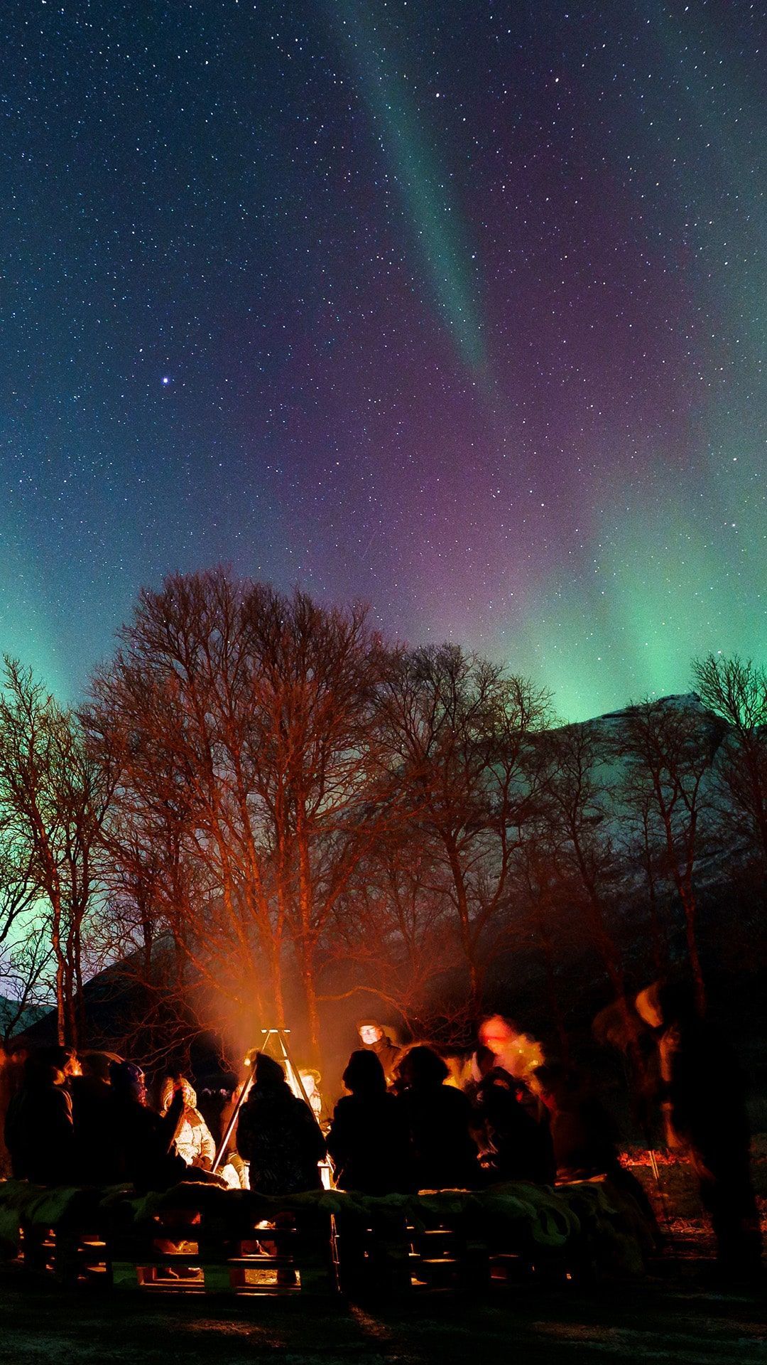 People around a campfire under the vibrant aurora borealis, with a starry sky.