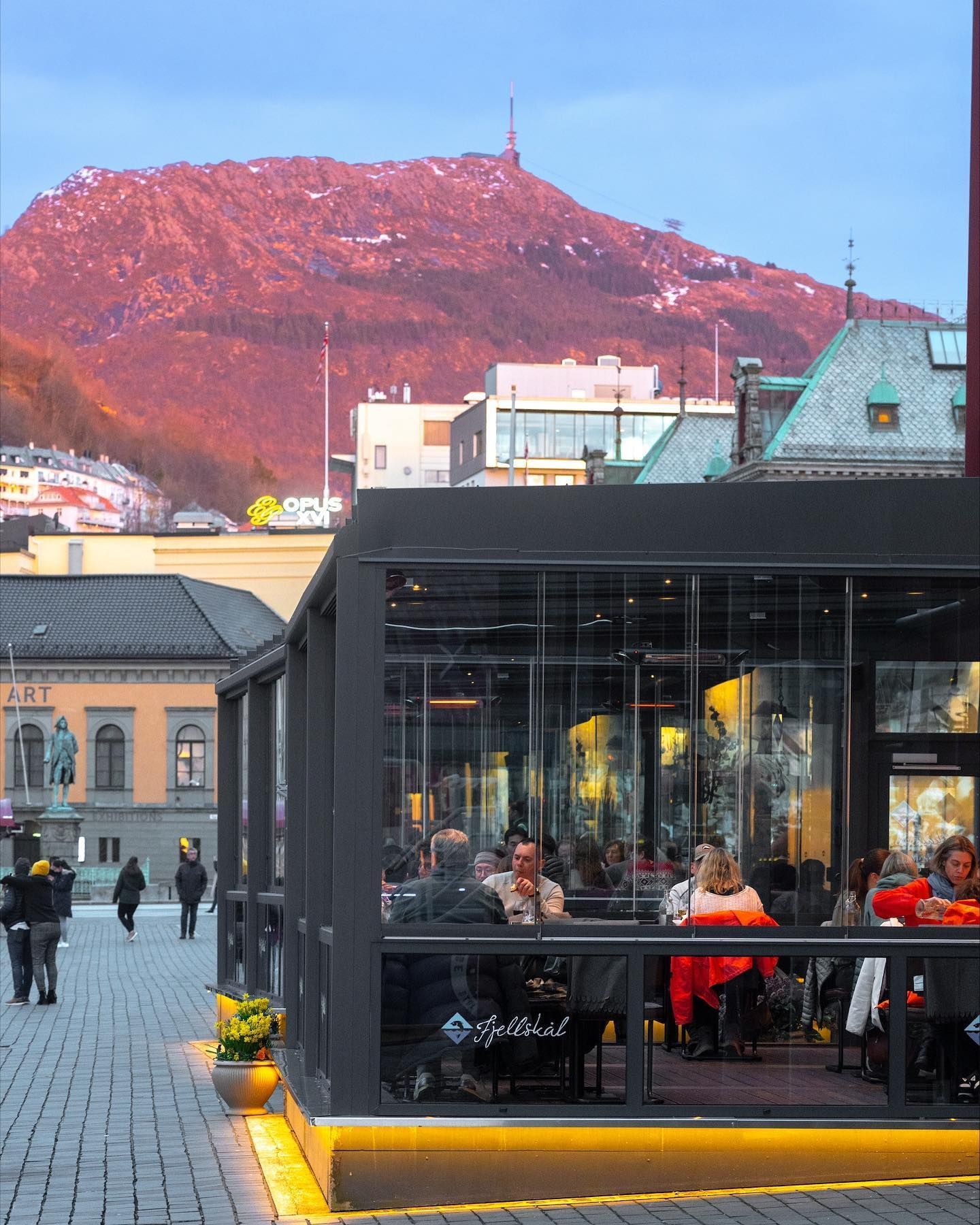Outdoor cafe with people, building, mountain in background at sunset.