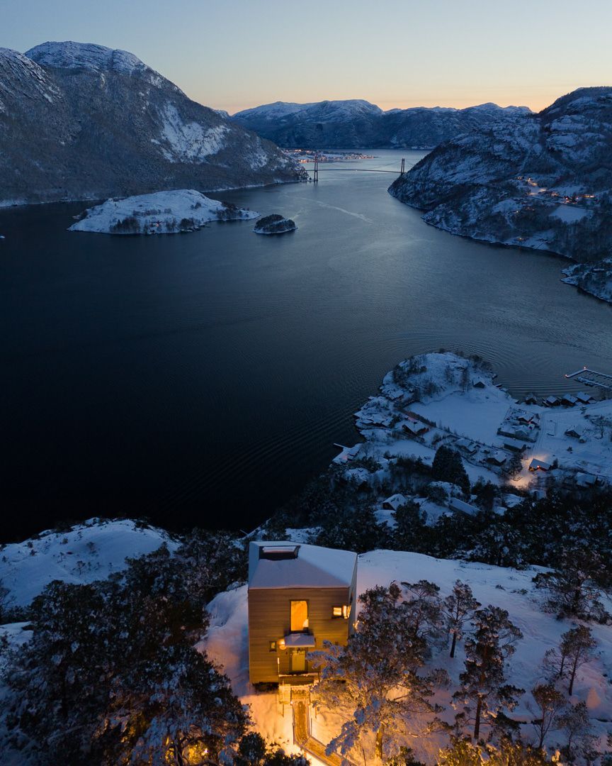 Snowy landscape with a modern house overlooking a dark fjord. Mountains in the background, lit windows in the house.