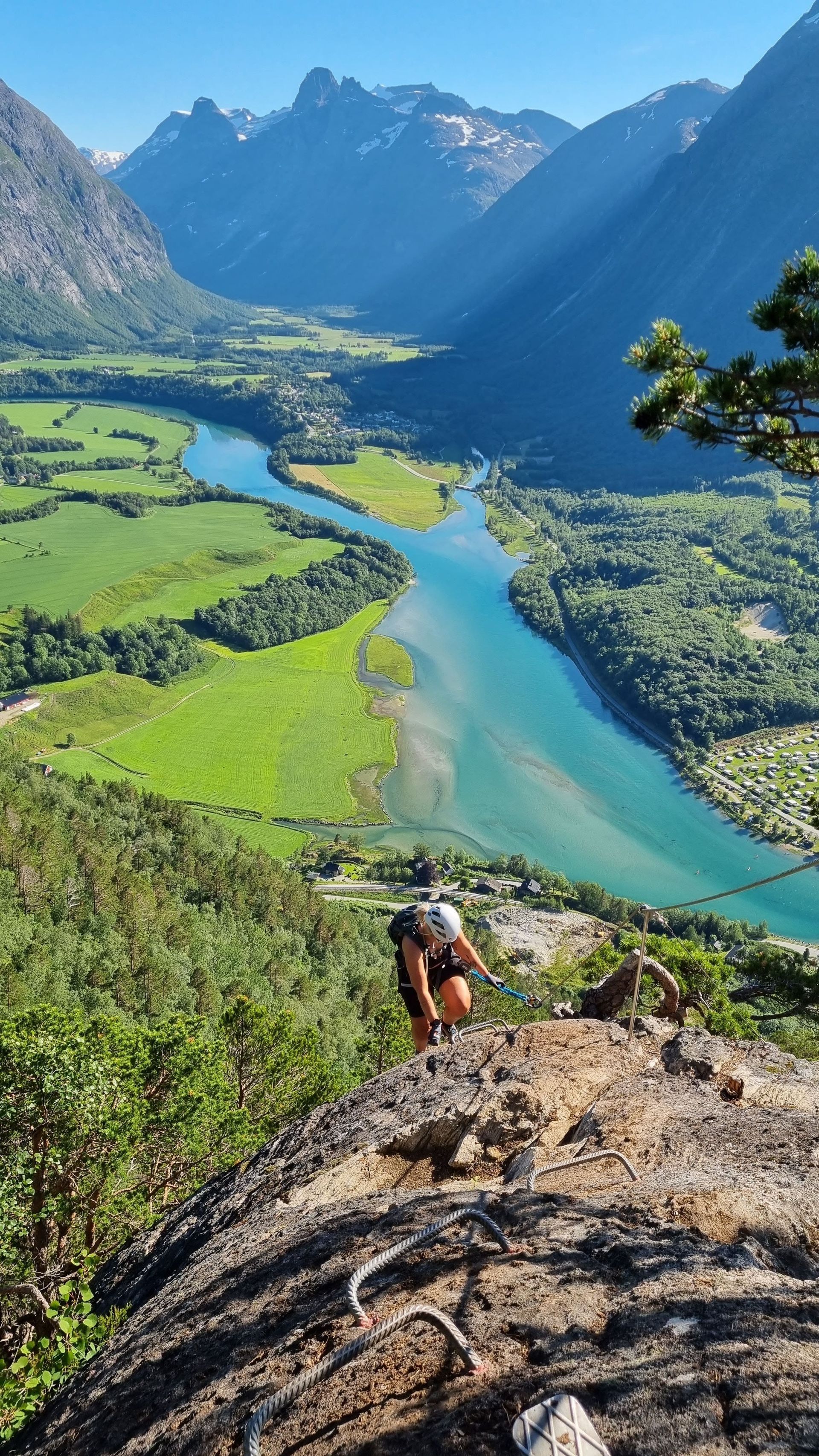 Person hiking up a rocky mountain trail overlooking a river valley with lush green fields and blue water under a bright sky.