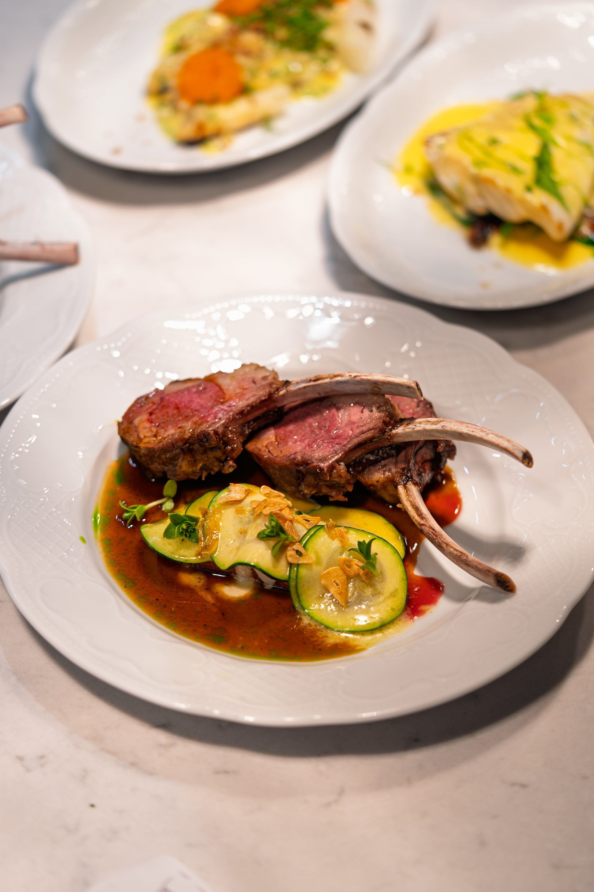 Rack of lamb with zucchini on a white plate, other dishes in background.