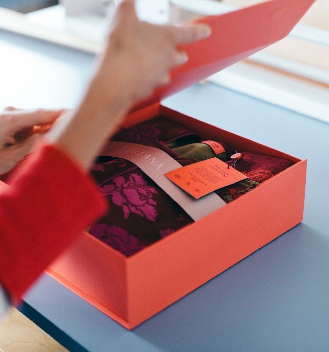 Hands opening a red box revealing a patterned scarf and tag inside.