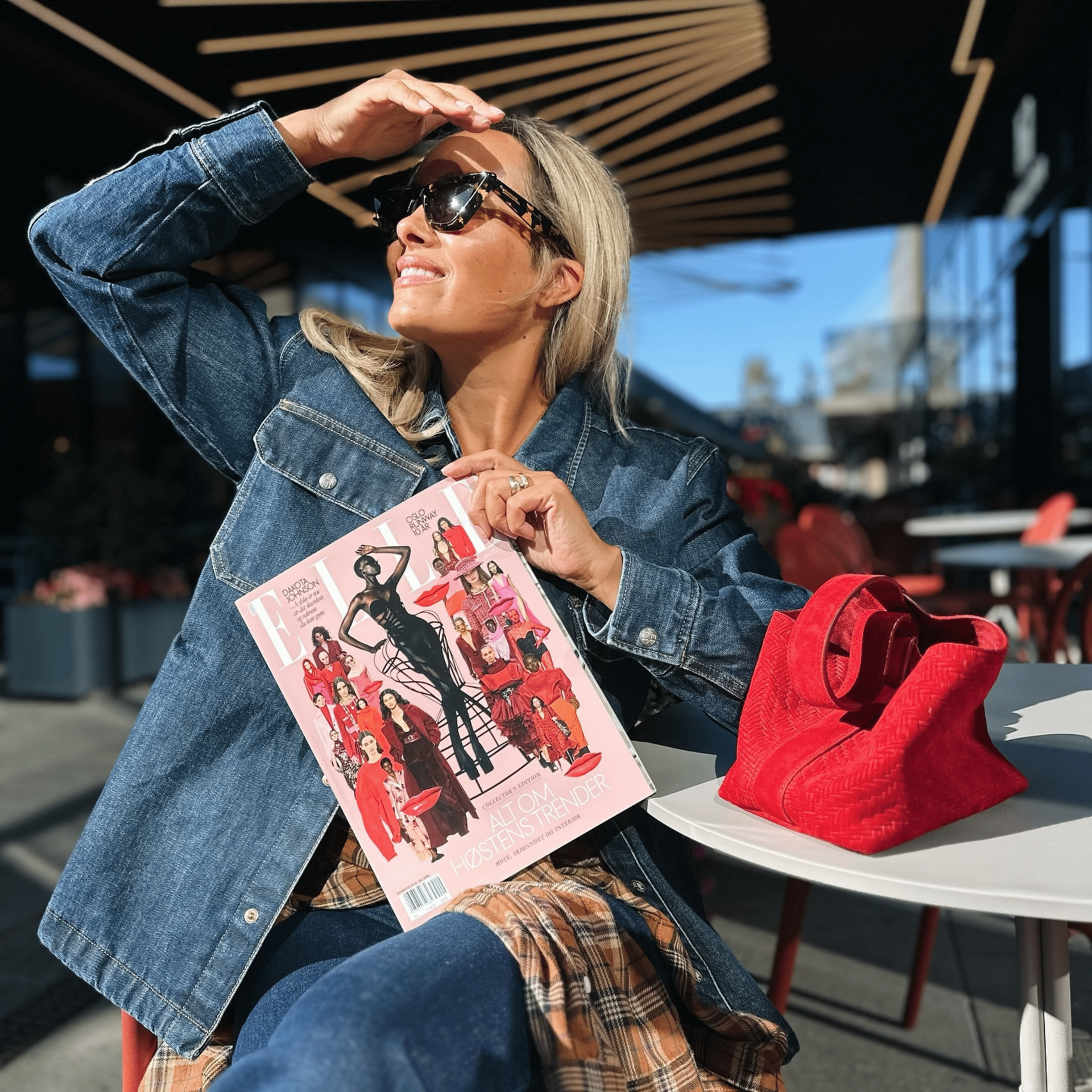 Woman in sunglasses shields her eyes, holding a magazine with a red bag on a cafe table.