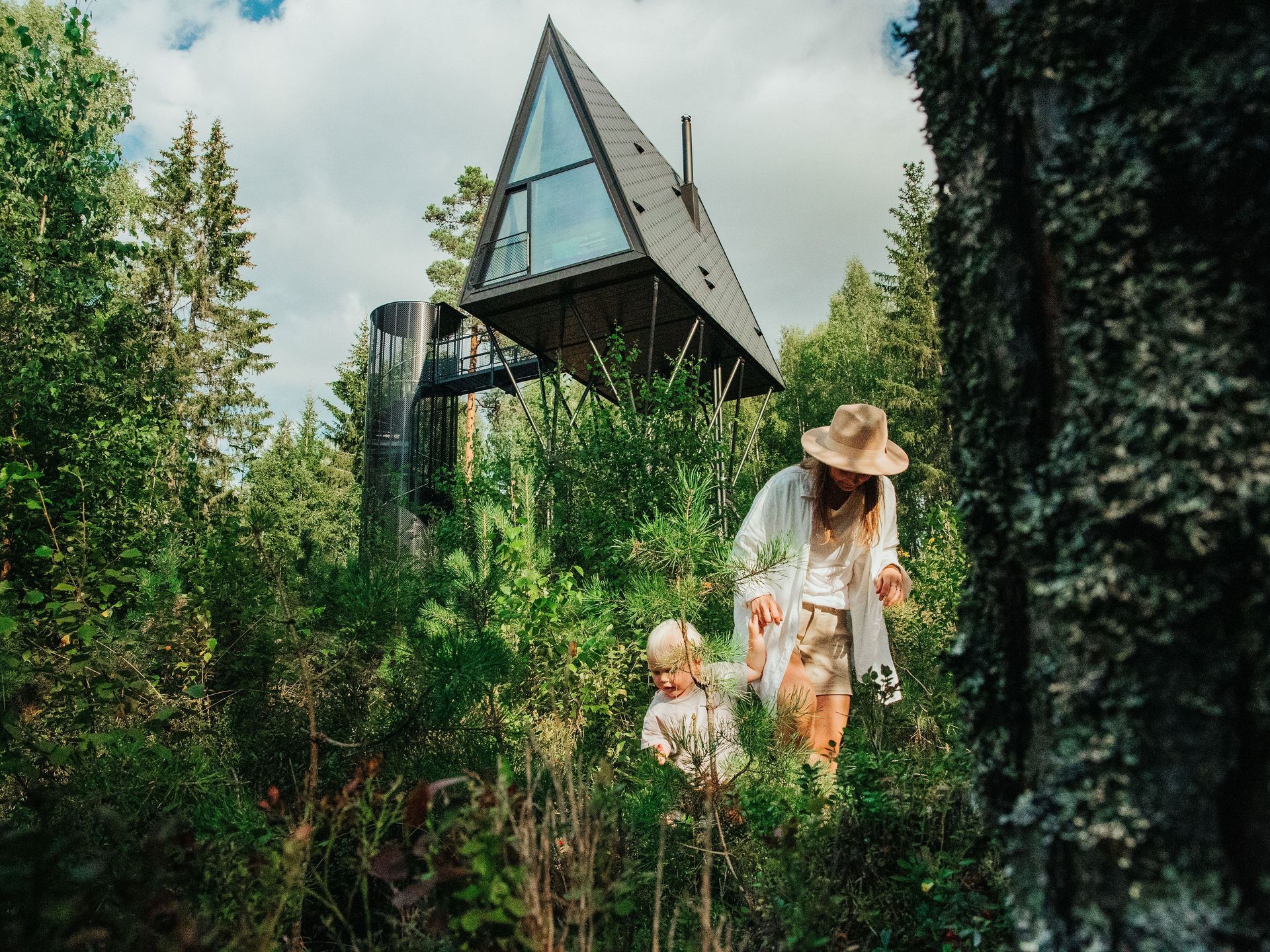 A-frame cabin in forest; person and child walking in brush.