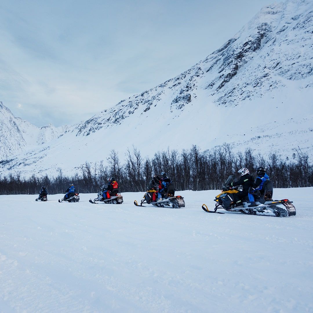 Snowmobiles travel across a snowy expanse near snow-covered mountains; riders are visible on the machines.