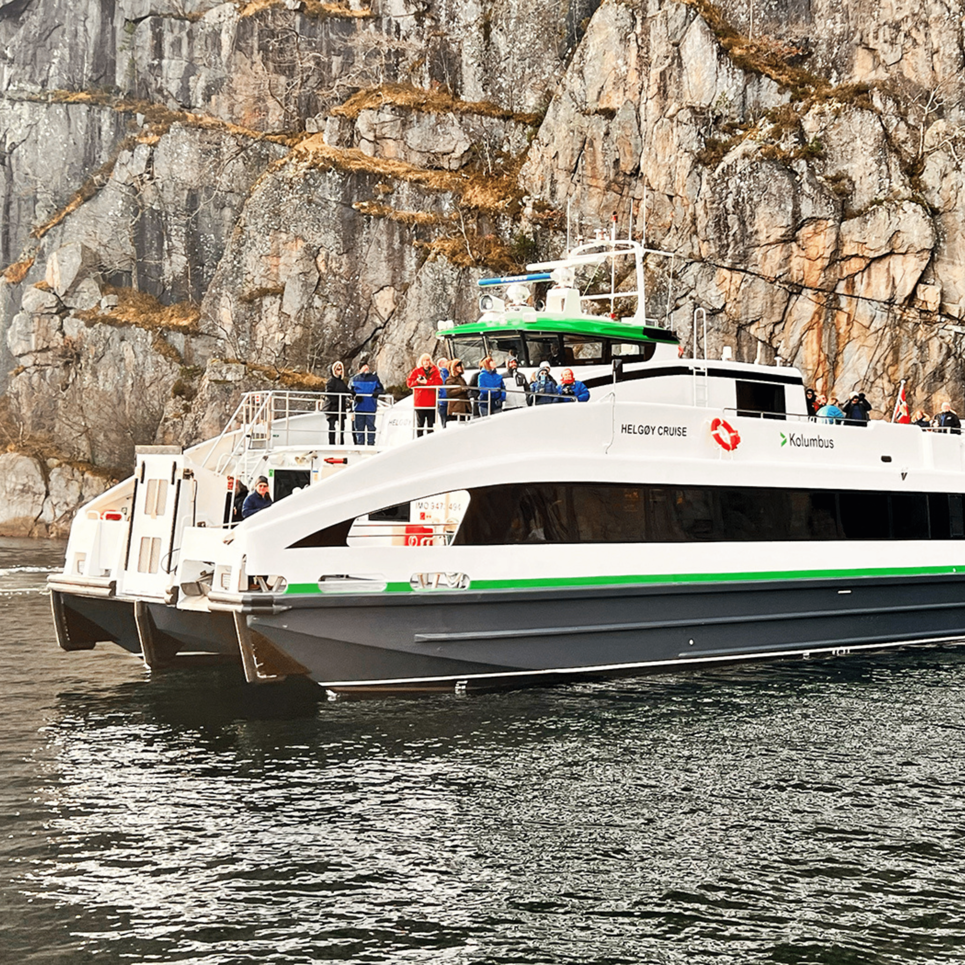 White and green catamaran ferry with passengers against a rocky cliffside.