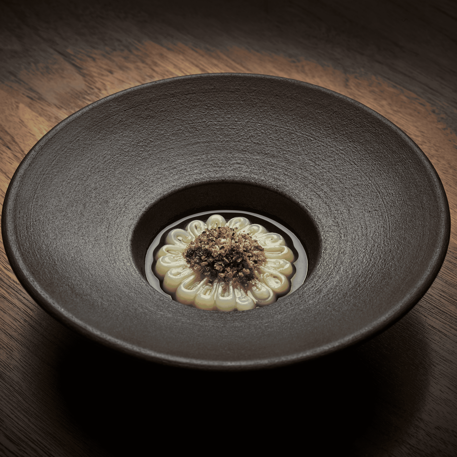 Dark bowl with a creamy, flower-shaped dish topped with dark crumbles in a pool of broth.