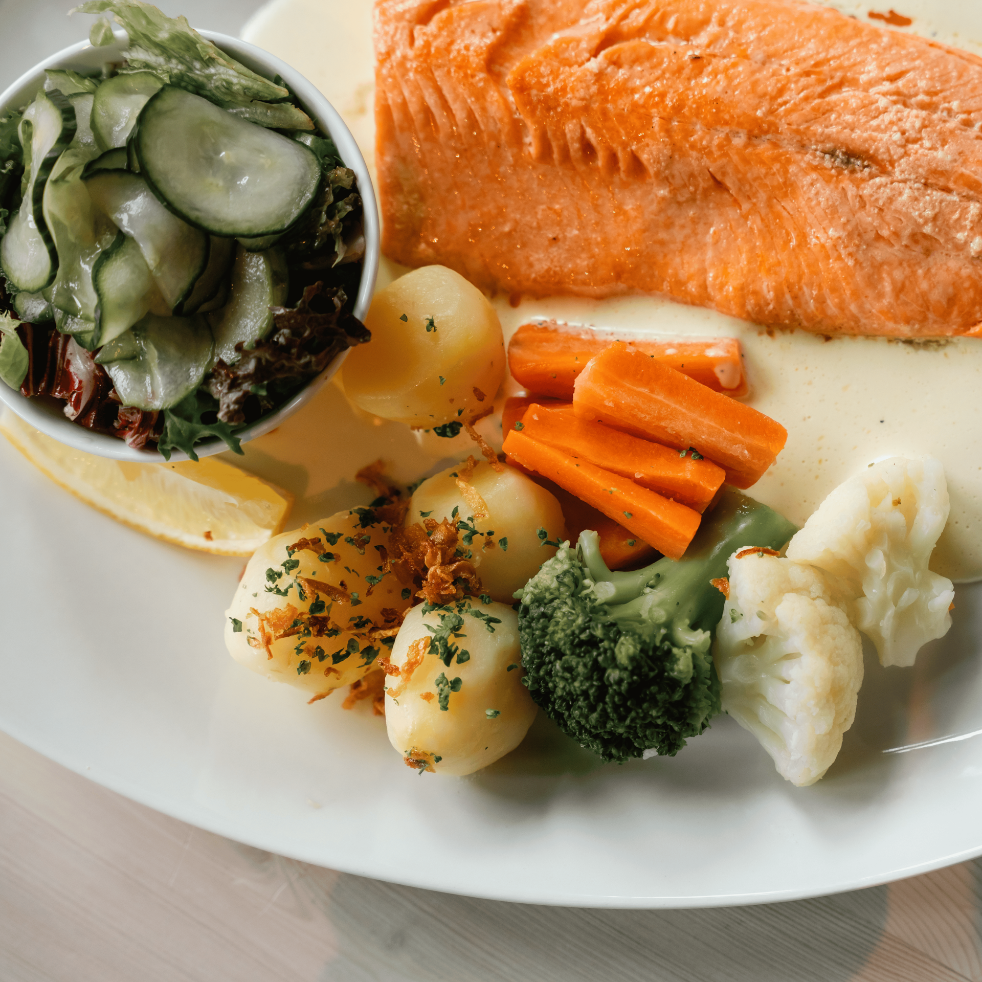 Salmon fillet, vegetables (carrots, broccoli, cauliflower, potatoes), salad, lemon wedge on a white plate.
