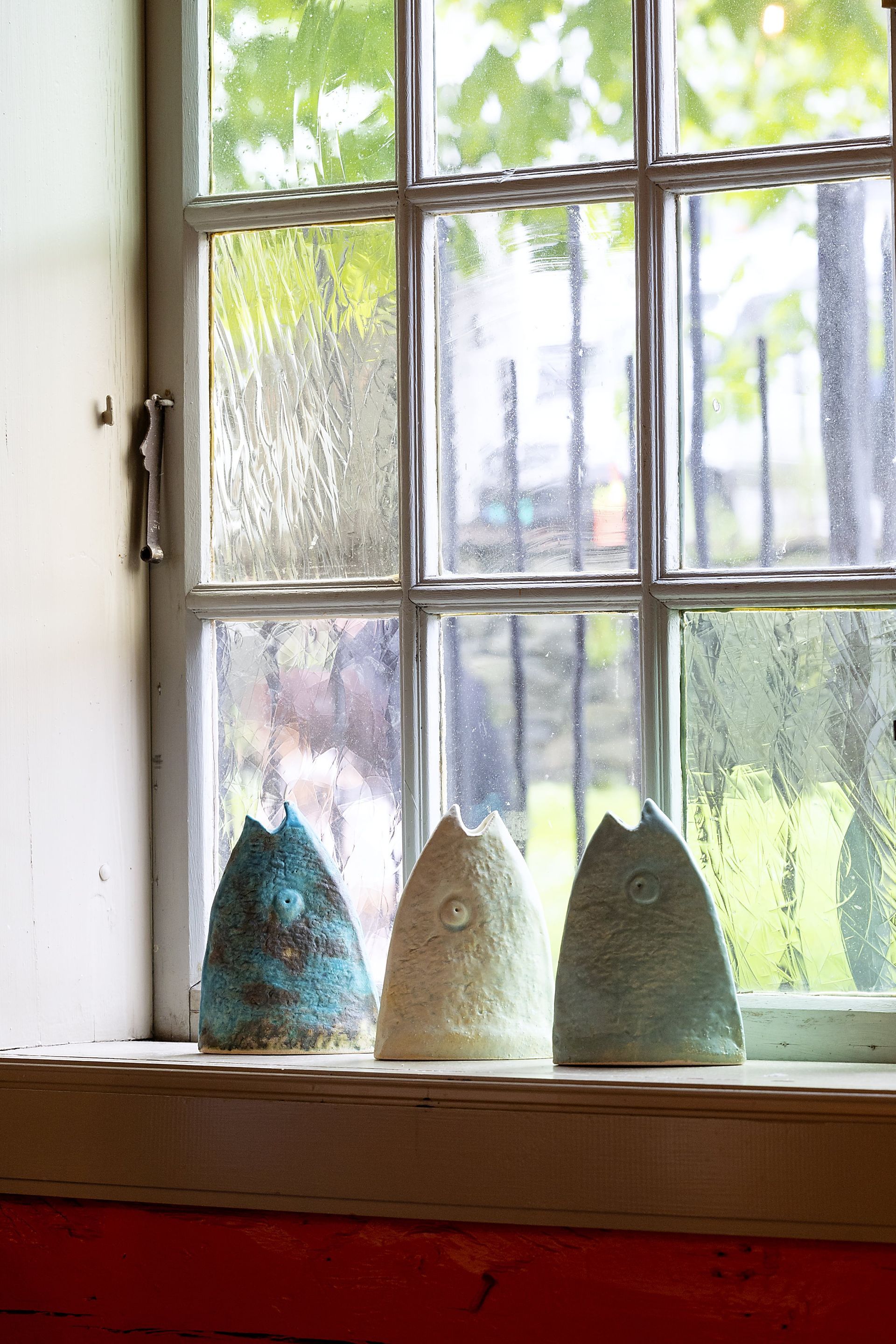 Three ceramic sculptures on a windowsill; blue, white, and green with textured surfaces, viewed against a rain-streaked window.