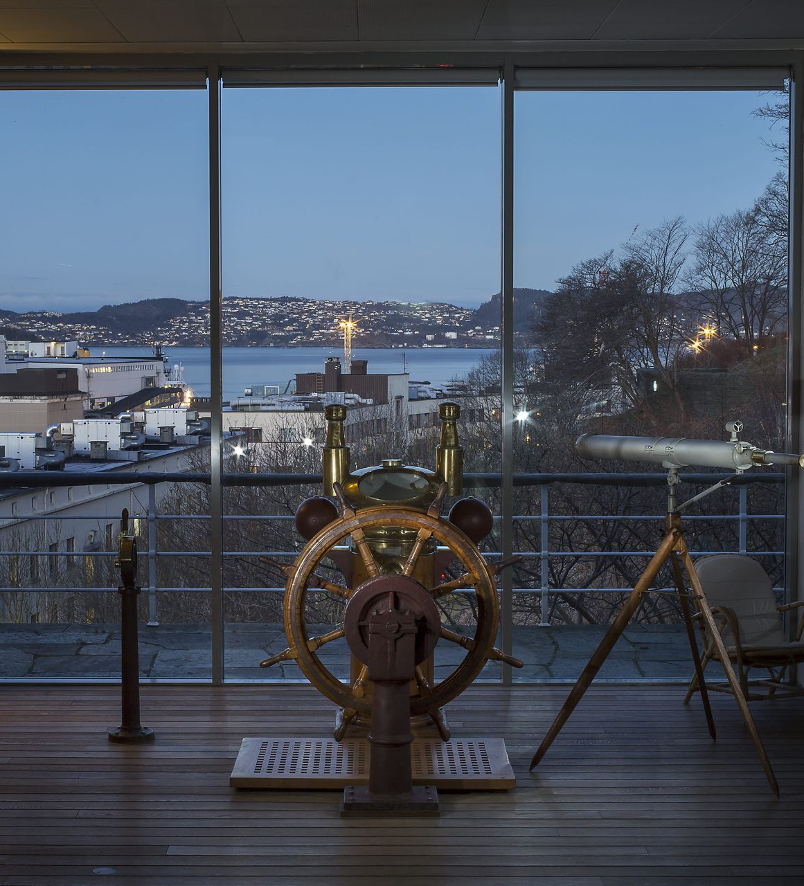 An interior view with a ship's wheel, telescope, and a window overlooking a cityscape at dusk.