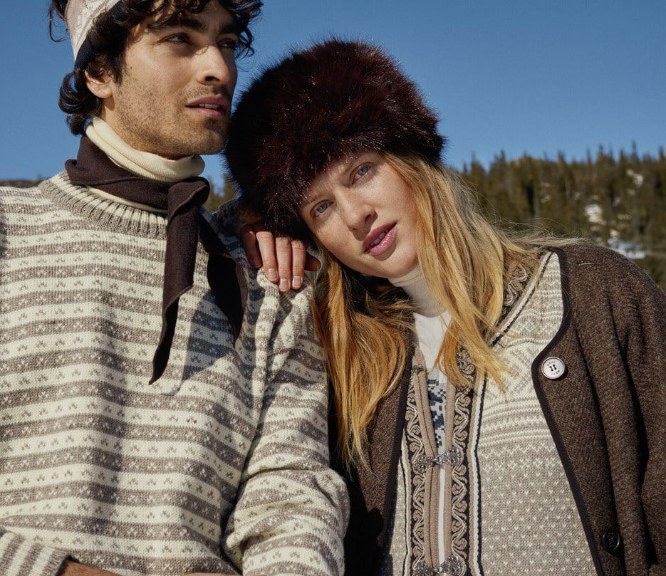 Couple in winter wear, man with sweater and scarf, woman with fur hat, looking at the camera outdoors.