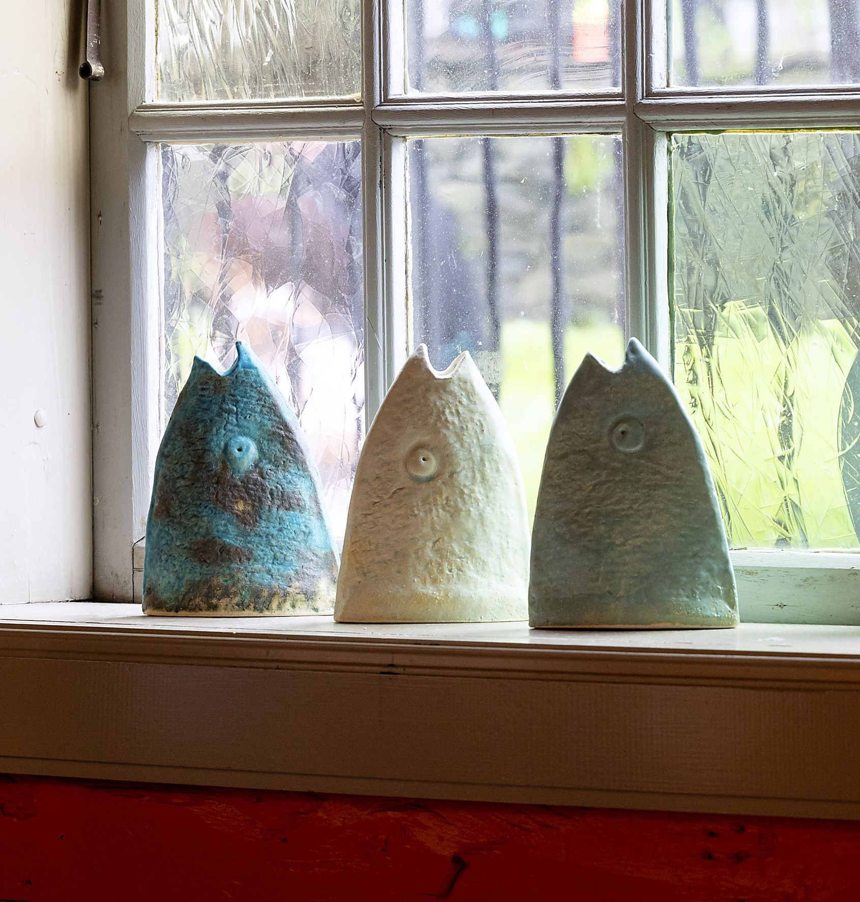 Three ceramic sculptures on a windowsill; blue, white, and green with textured surfaces, viewed against a rain-streaked window.