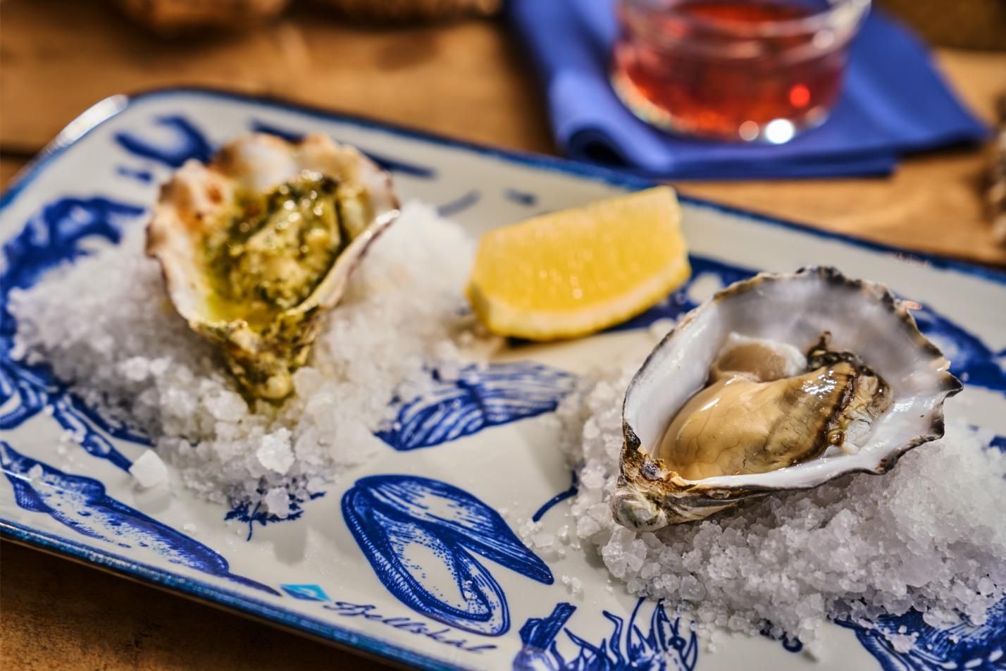 Oysters on a blue and white patterned plate with rock salt and lemon. One oyster is topped with green sauce.