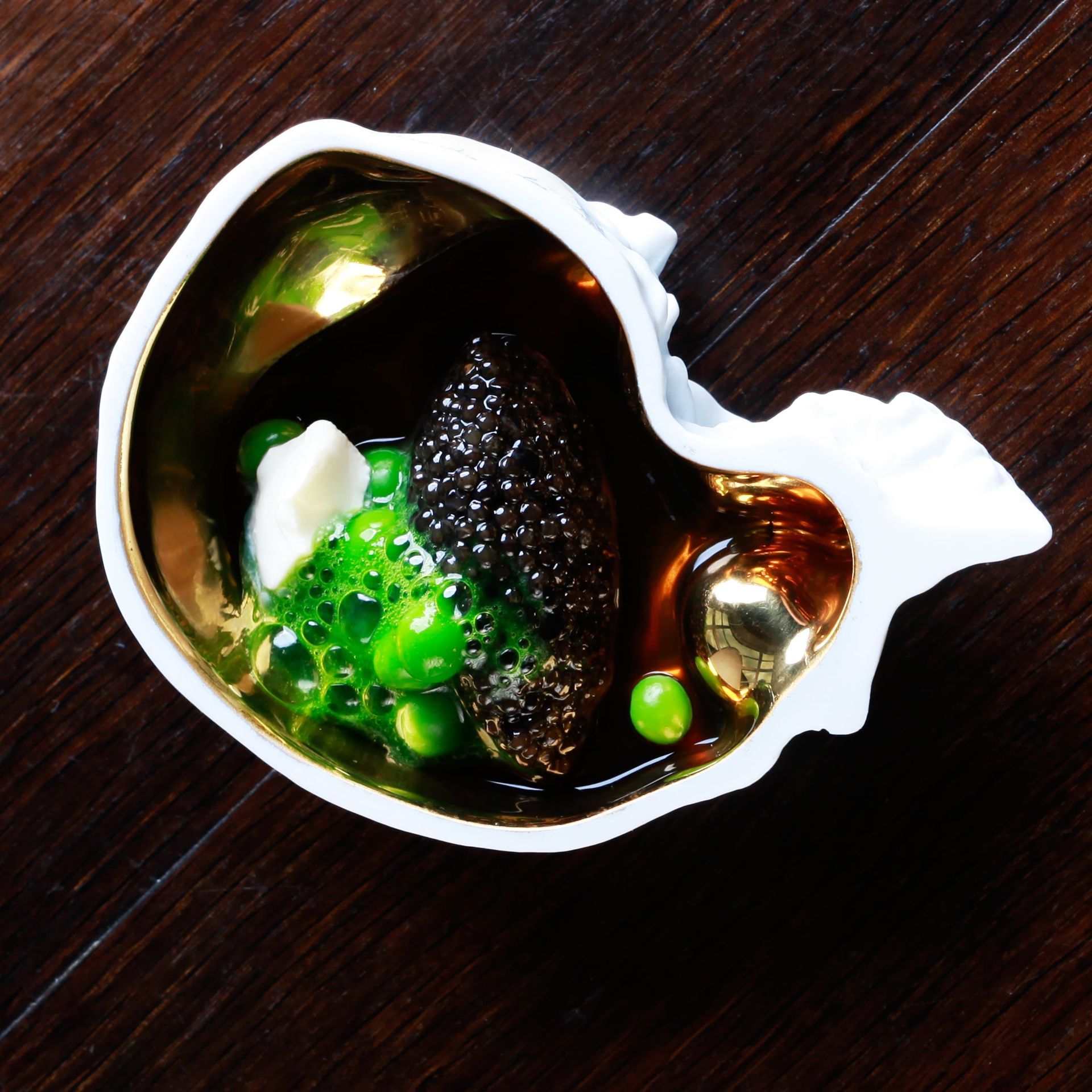 A small, shell-shaped bowl with gold interior, containing caviar, green peas, and sauce, on a wood table.