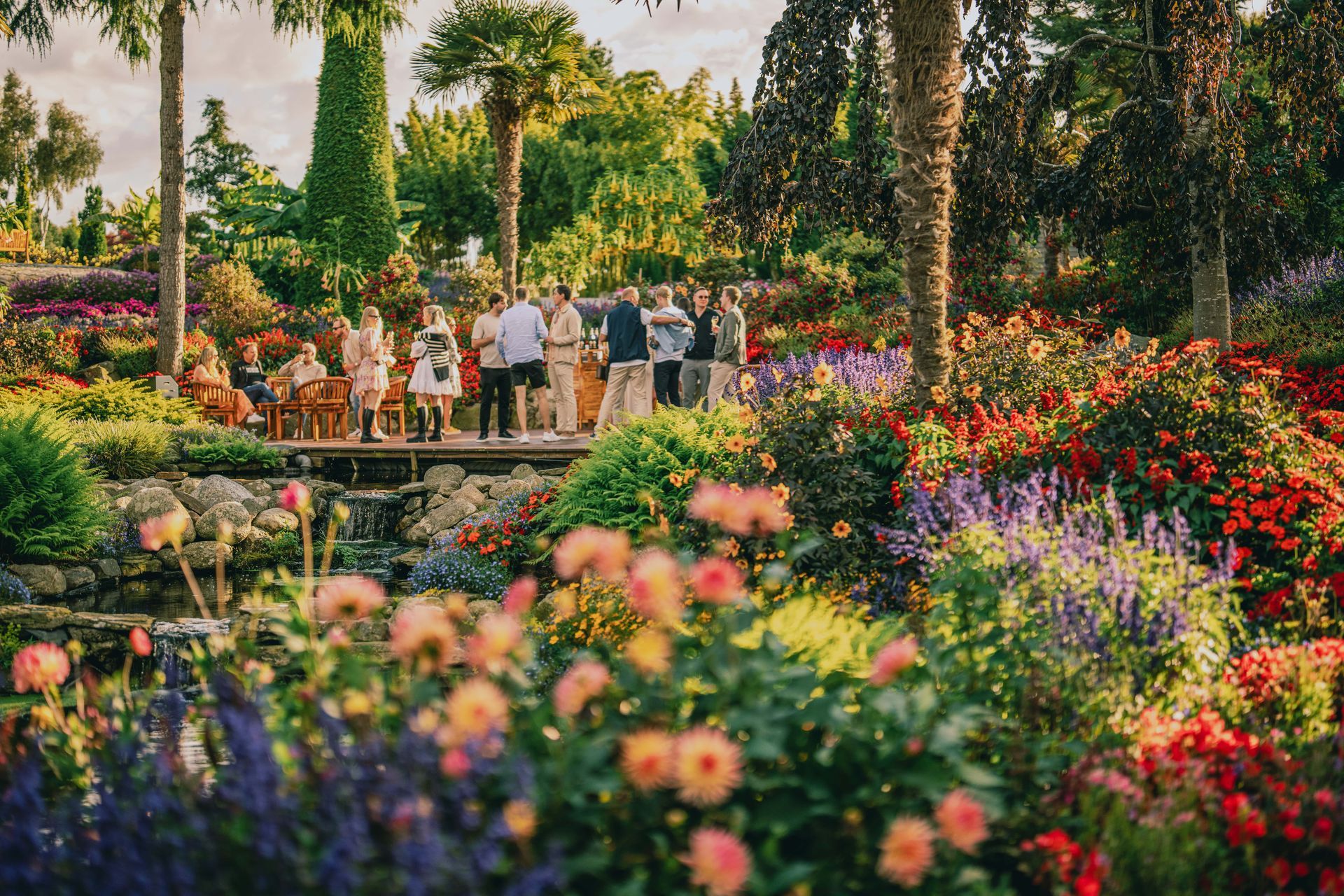 Group of people on a bridge in a colorful garden, flowers in bloom under a sunny sky.