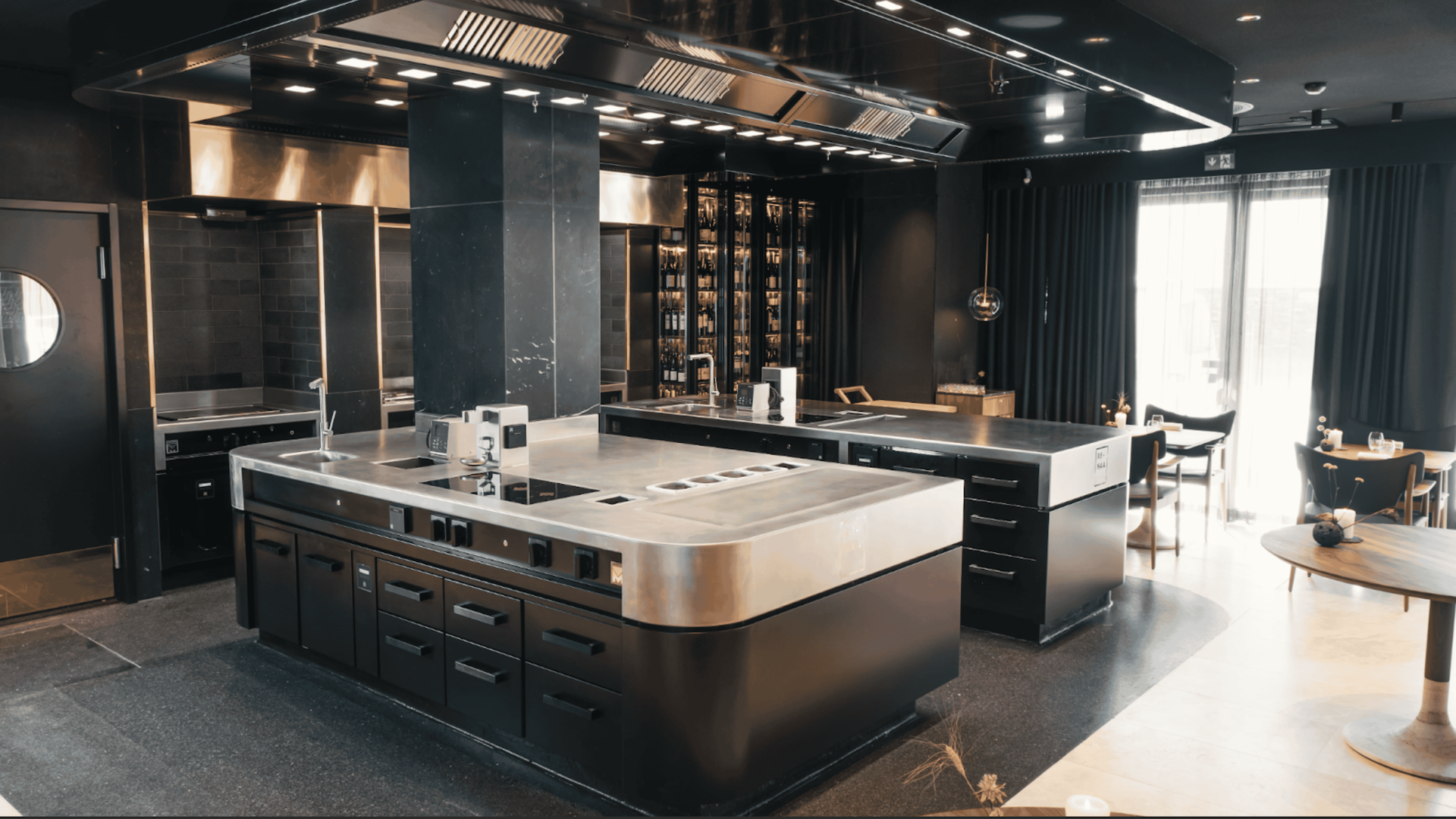 Modern, dark kitchen with stainless steel appliances, central island, and dining area with curtains.