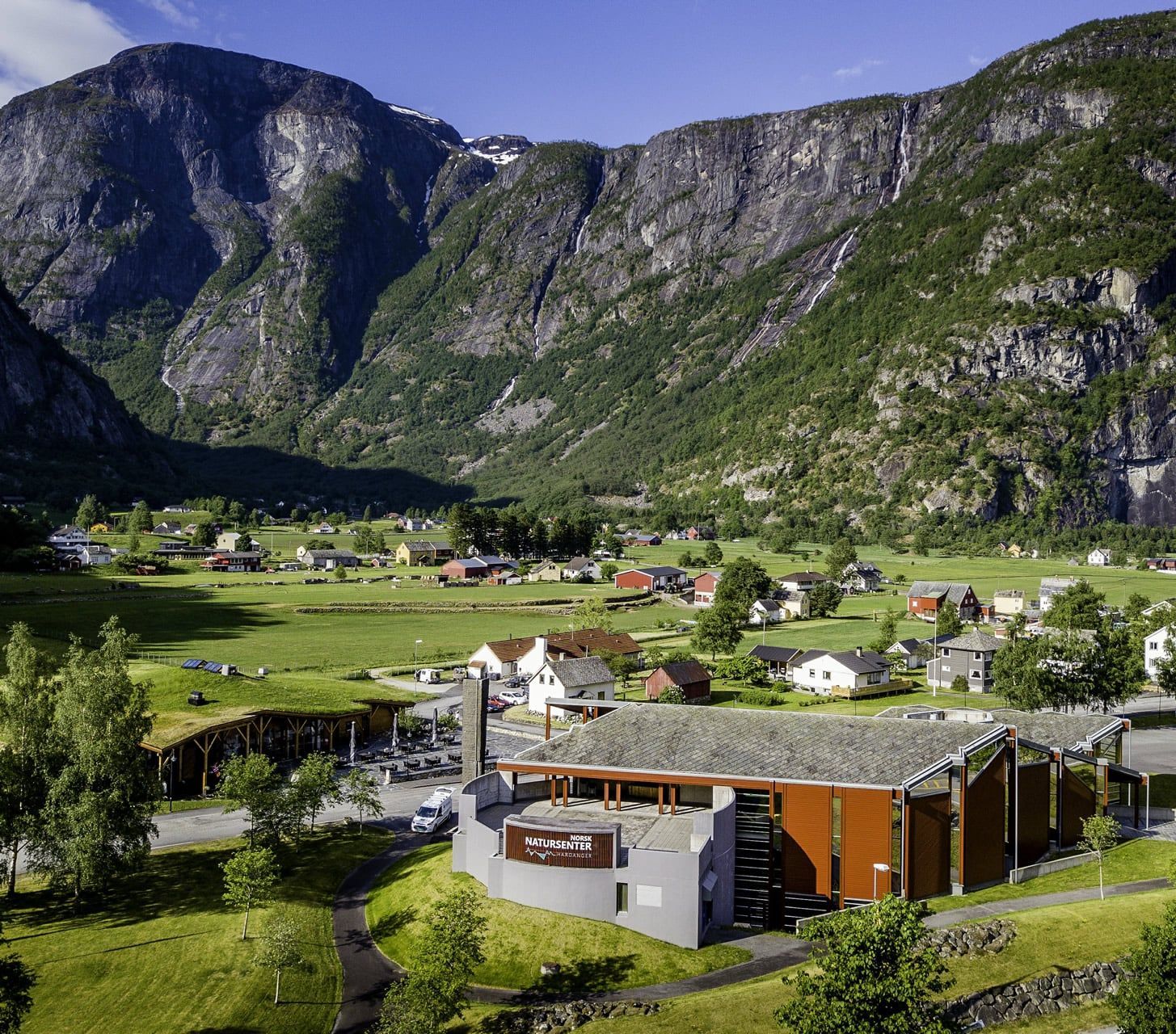 A village nestled between green fields and towering mountains with a modern building in the foreground.