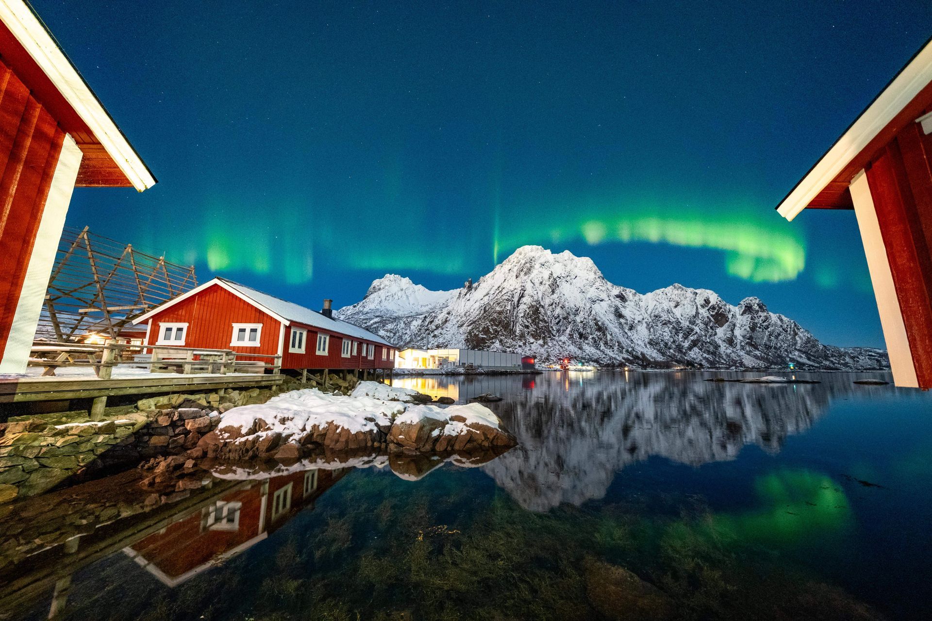 Red cabins by water, snow-capped mountains and green Northern Lights in the sky at night.