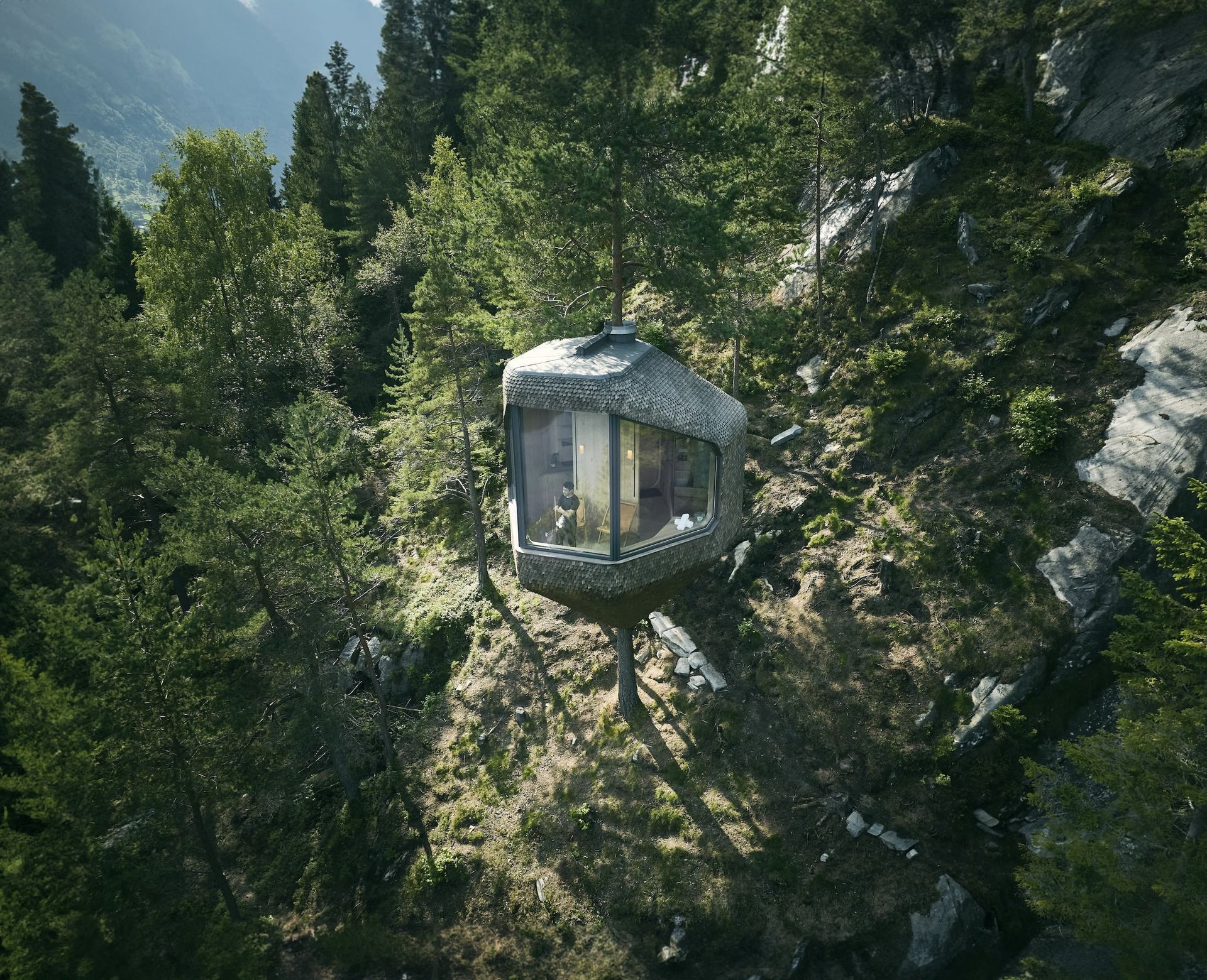 Drone view of a stone-clad cabin nestled in a hillside forest with a large window facing the viewer.