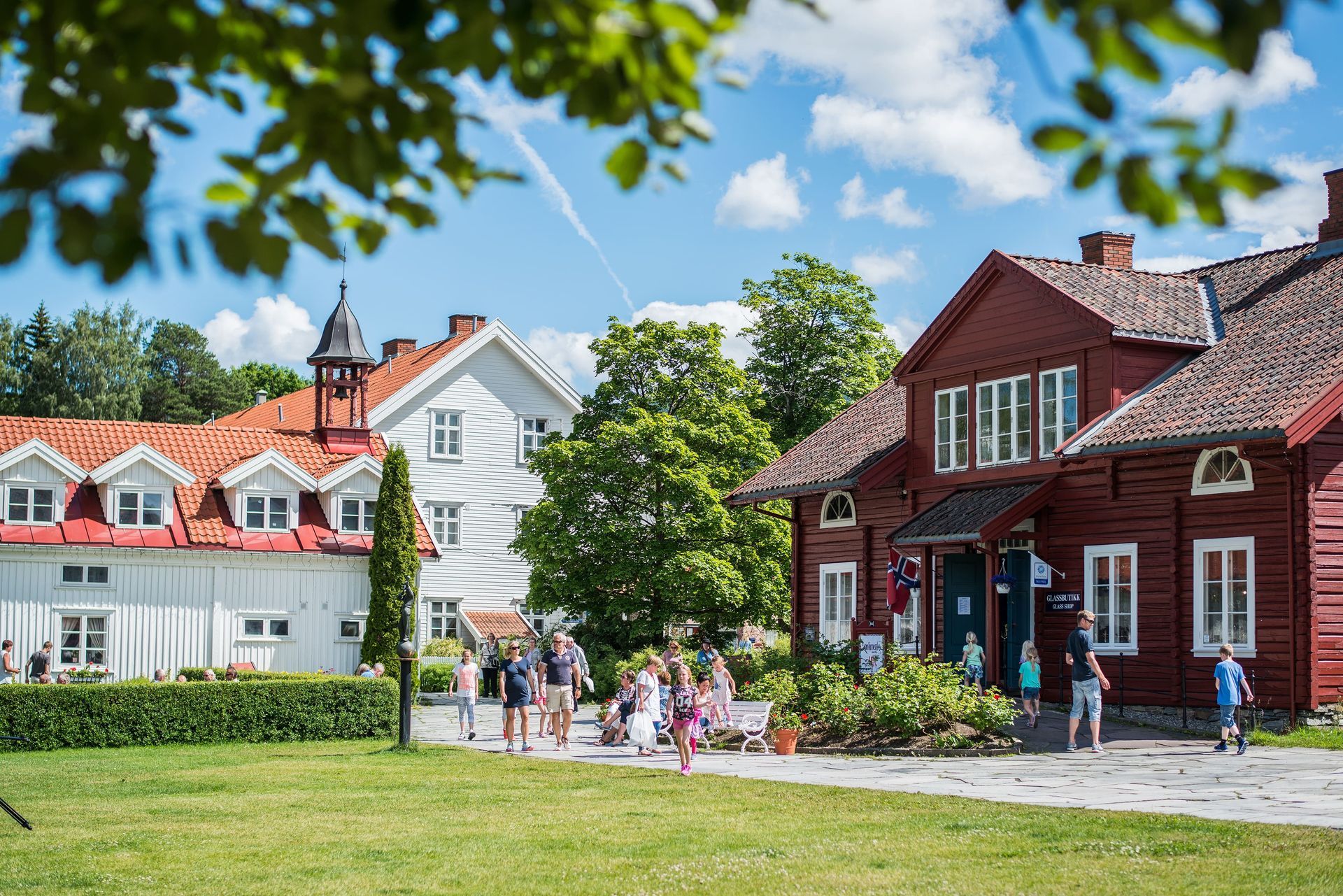 Red and white buildings with people outside on a sunny day. Green grass and trees frame the scene.