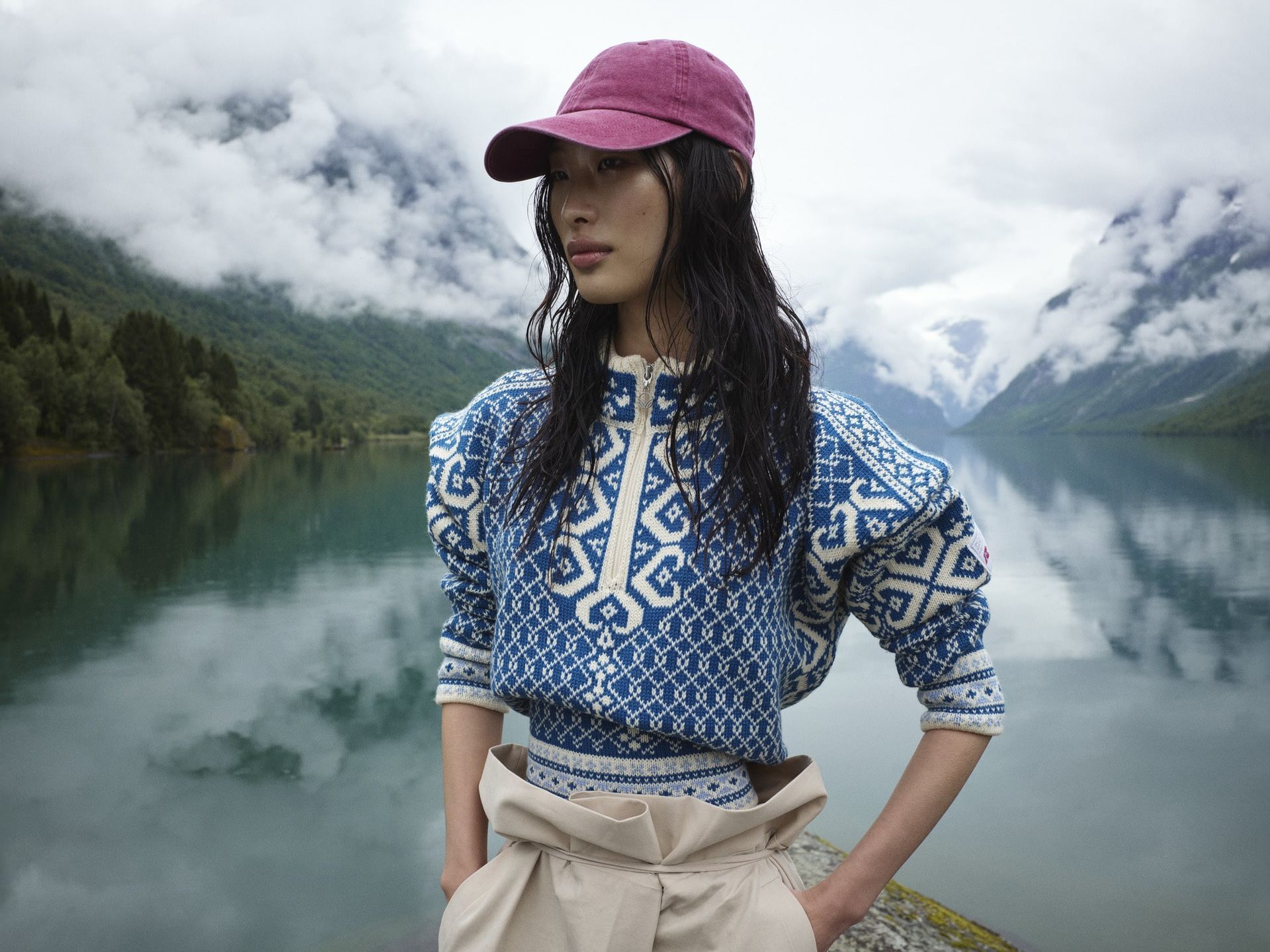 Woman in blue patterned sweater and maroon cap, standing by a lake with mountains in background.