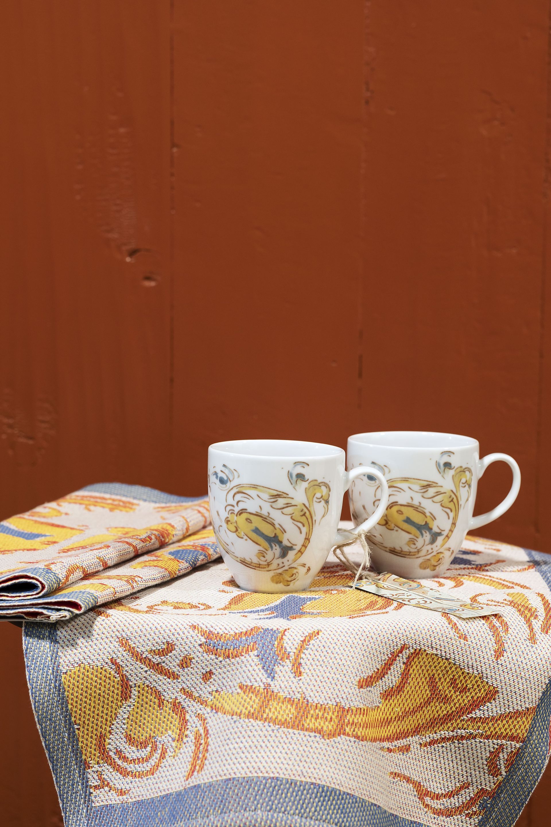Two white mugs with a floral design on a patterned cloth, against a brown wall.