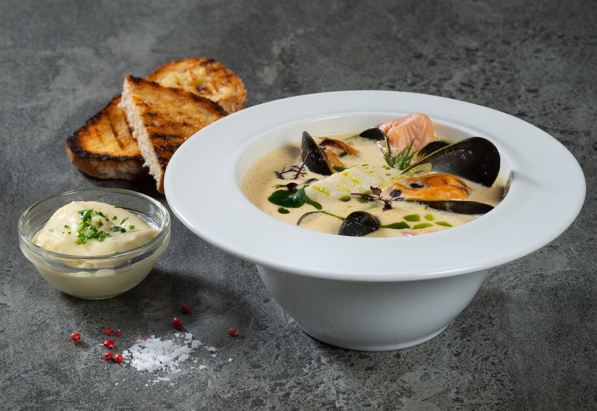 Creamy seafood stew in a white bowl with toasted bread and dip. Grey background with salt and peppercorns.