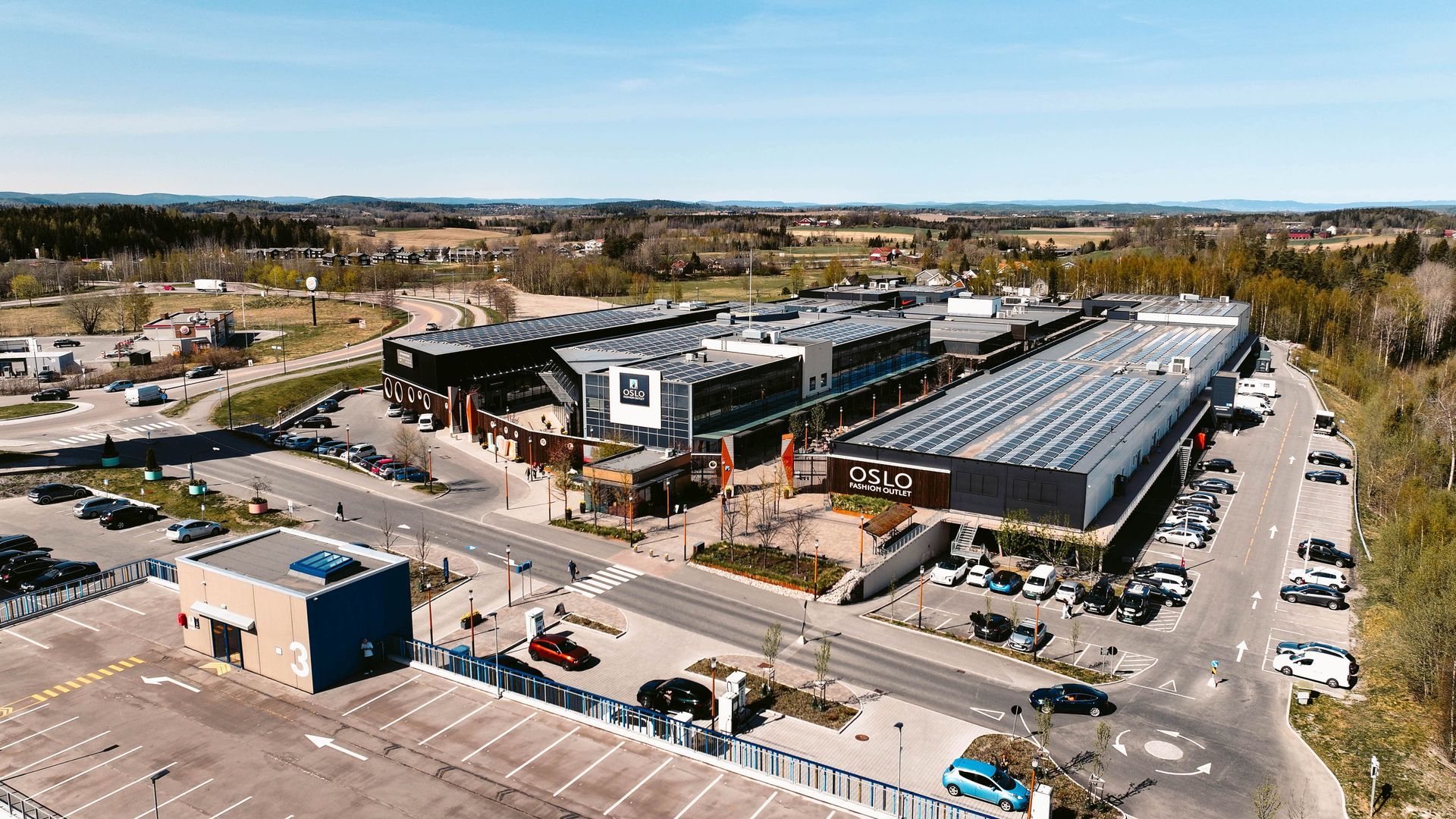 Aerial view of a modern shopping center with various stores, surrounded by parking and roads.