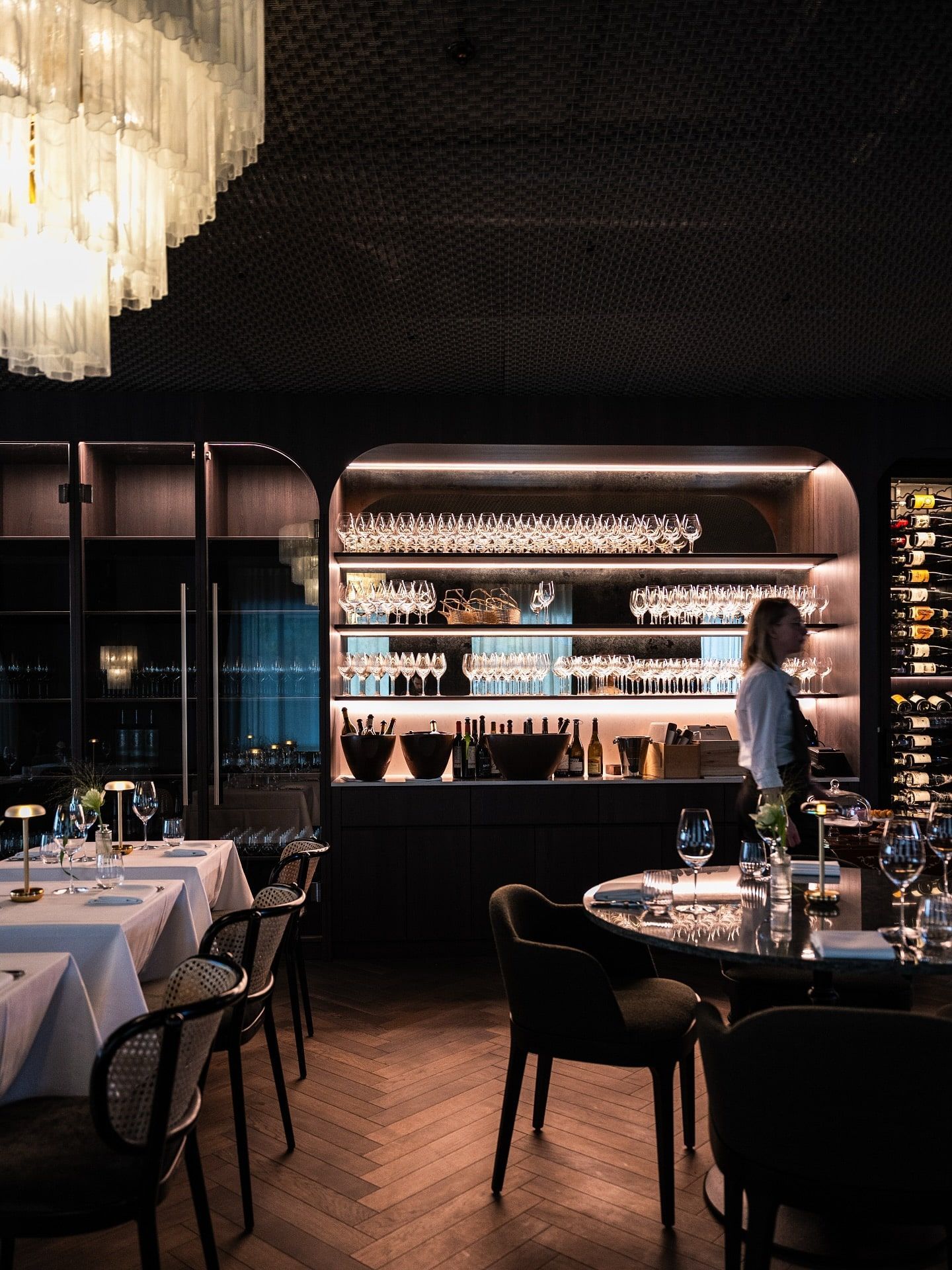 Elegant restaurant interior with tables set for dining; a person stands near a backlit bar with wine glasses.