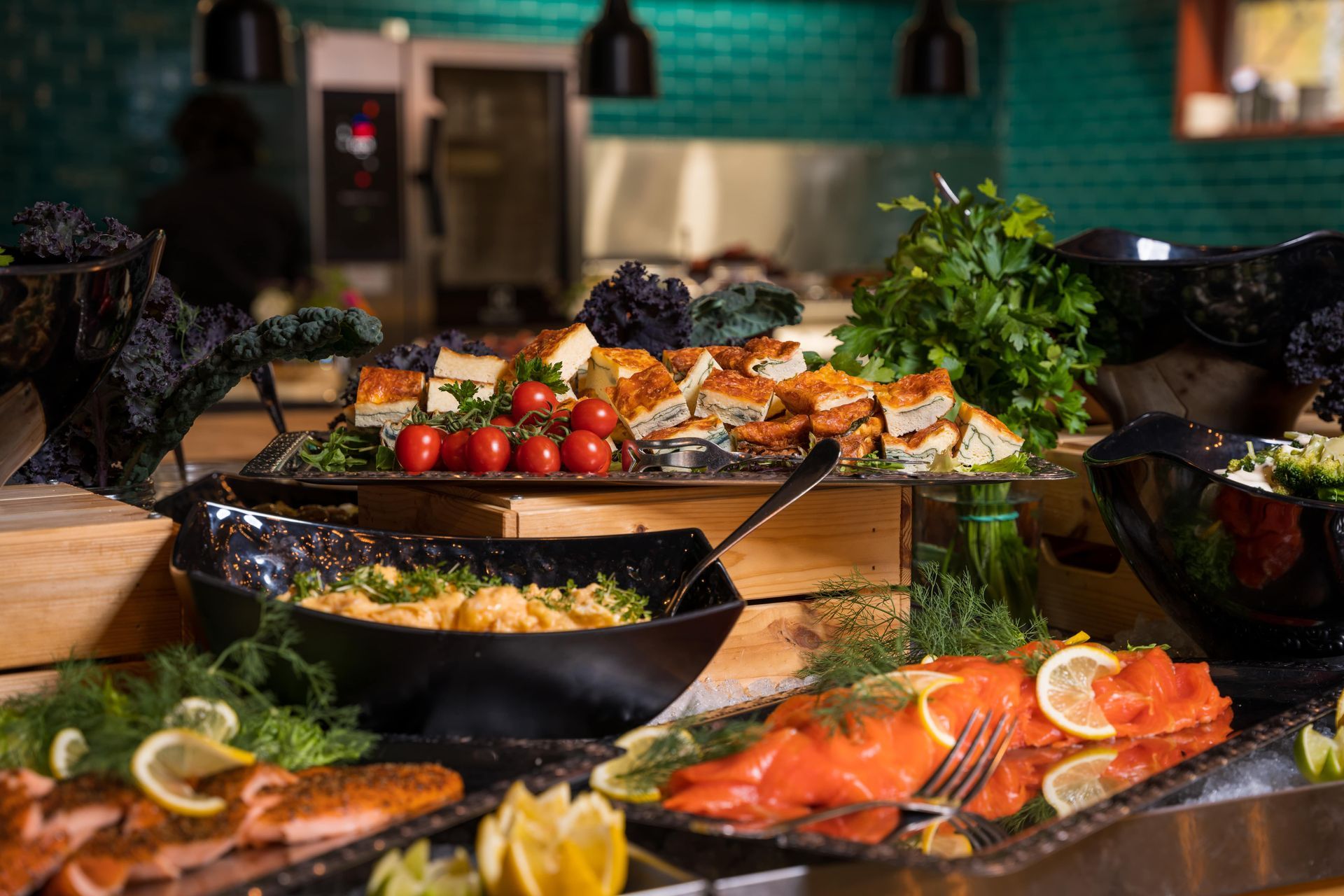 A buffet spread with various dishes like smoked salmon, pasta, tomatoes, and pastries, set against a teal-tiled wall.