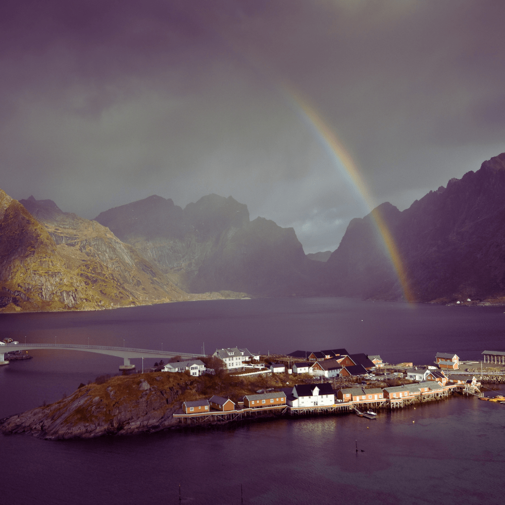 Island village under rainbow in fjord with mountains.