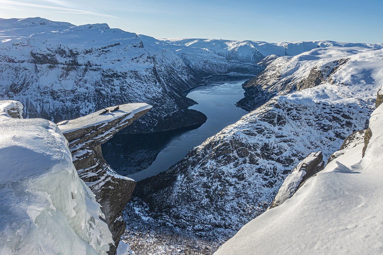 Snow-covered mountain range overlooking a long, dark lake under a clear, blue sky.