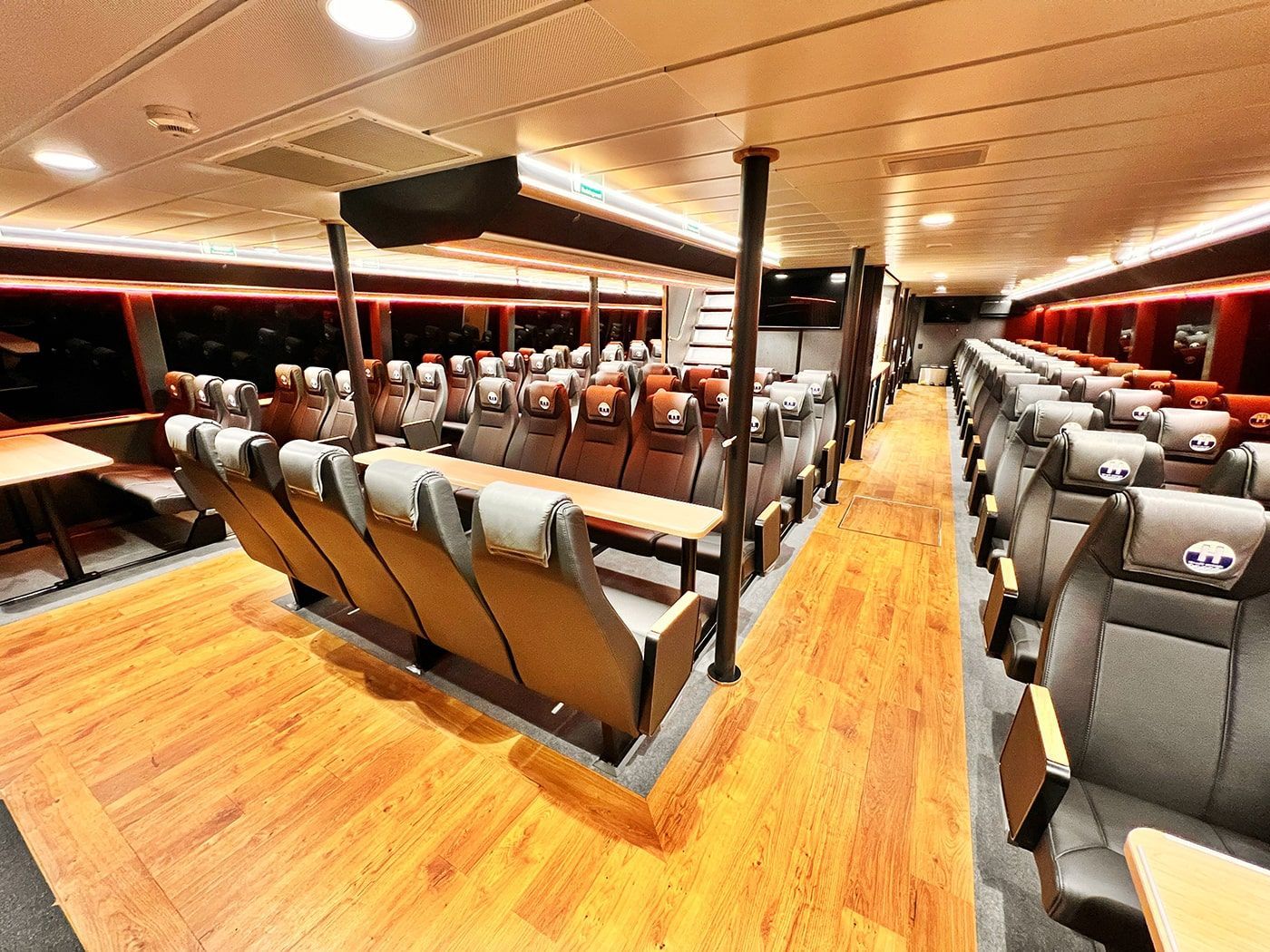 Interior of a ferry with rows of gray seats and wooden flooring. Red accents and windows.