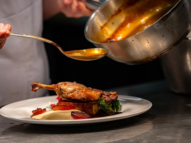 Chef pouring sauce over a plated dish of cooked meat and vegetables.