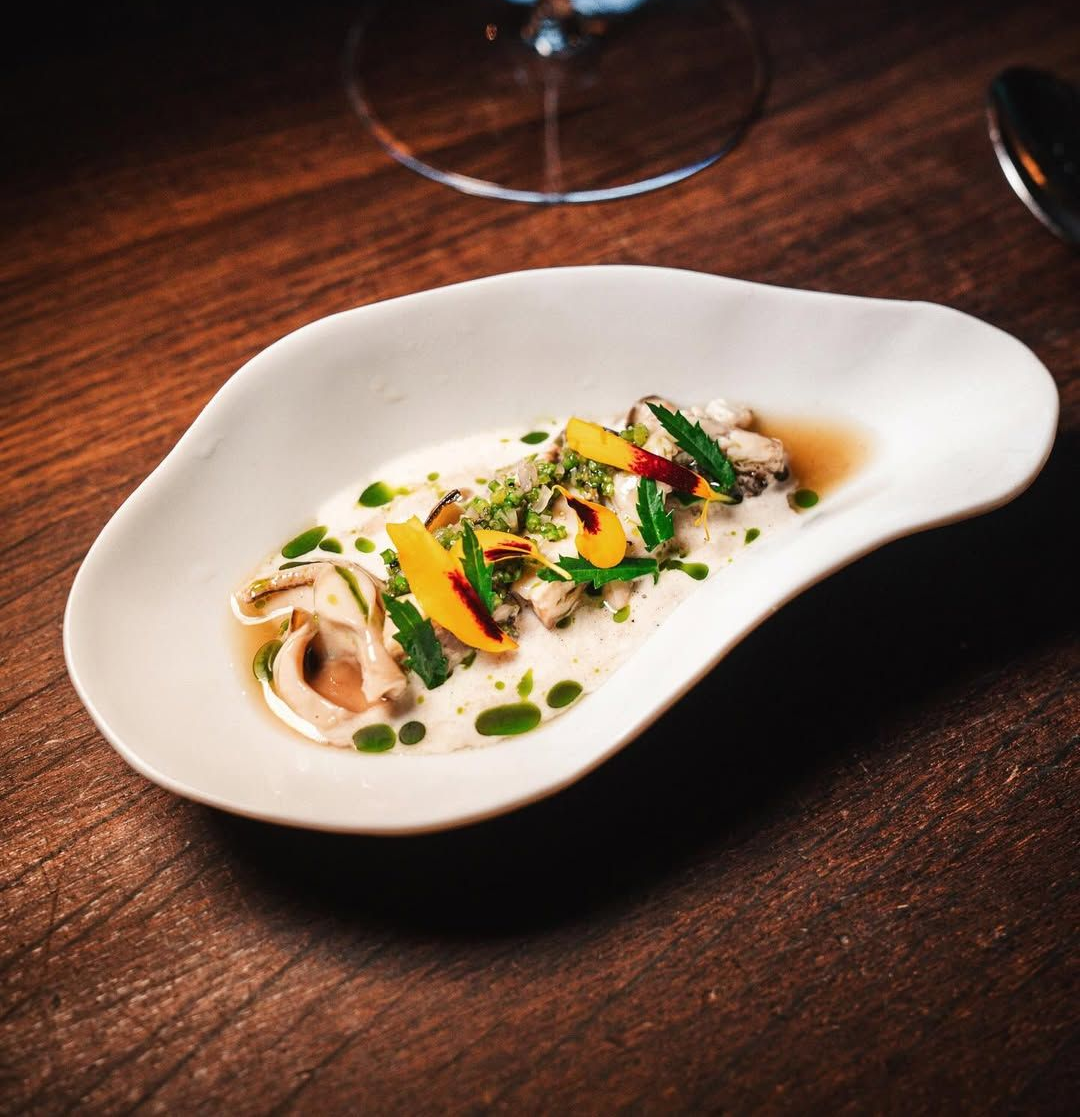 Appetizer on an organic-shaped white plate: shellfish, herbs, and sauces on a dark wood table.