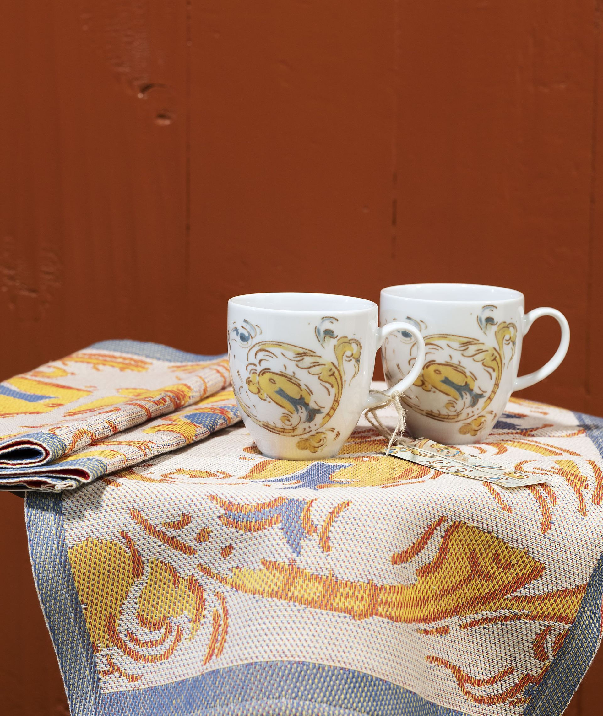 Two white mugs with a floral design on a patterned cloth, against a brown wall.