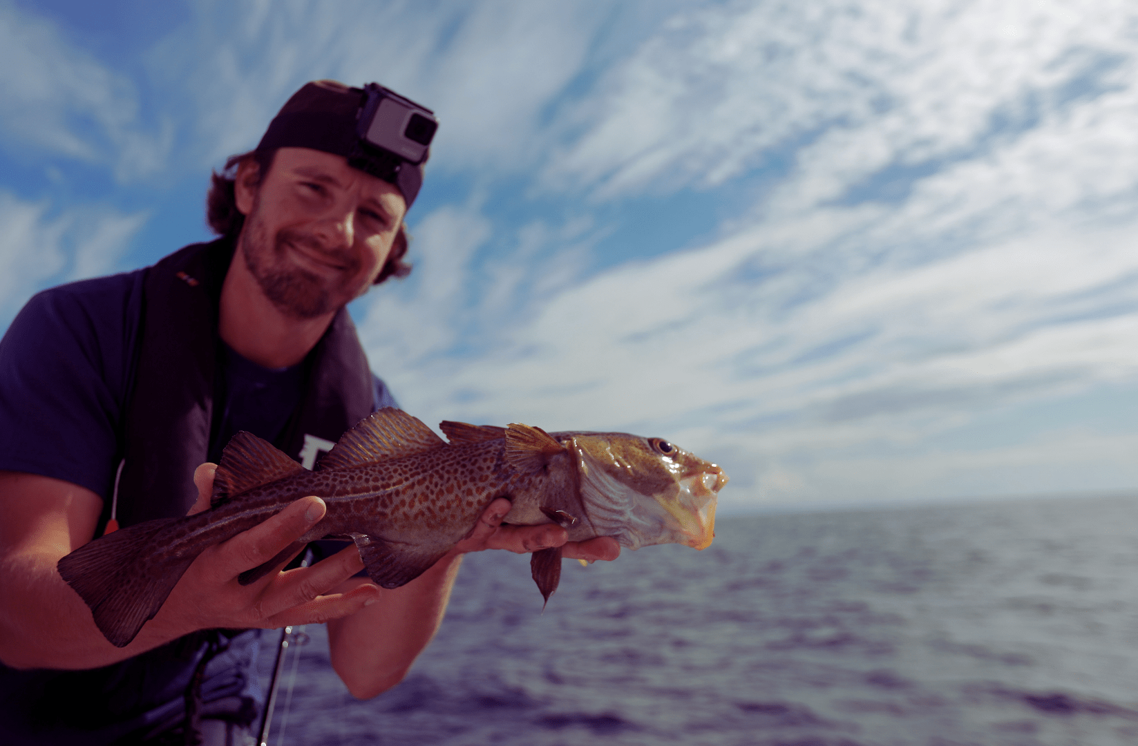 Man smiling, holding a large fish on a boat; ocean and cloudy sky in background.