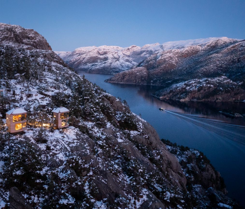 Two cabins nestled on a snowy cliffside overlooking a fjord in Norway.