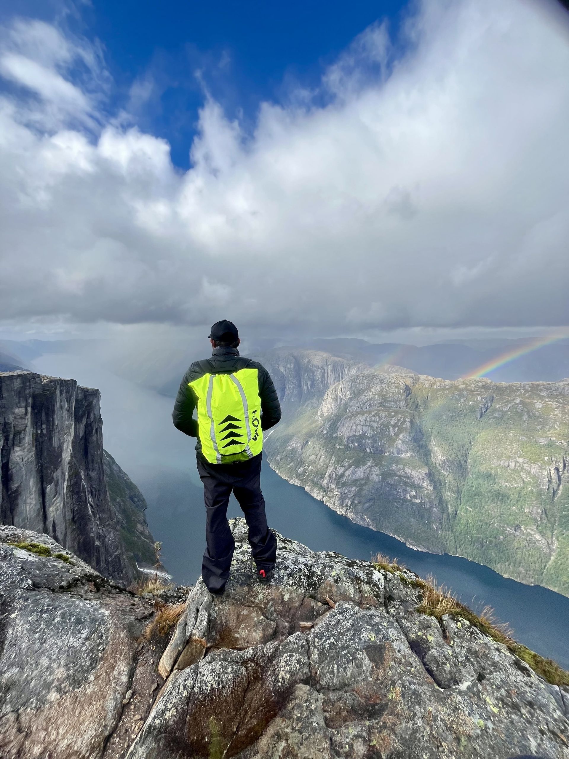 Person on a mountaintop, overlooking a fjord, with a rainbow. The person wears a neon green backpack and dark clothing.