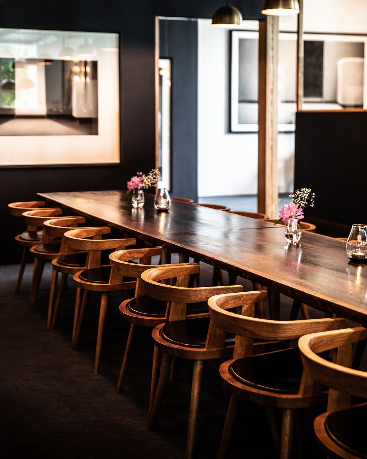 Long wooden table with curved wooden chairs in a dimly lit restaurant. Pink flowers adorn the table.